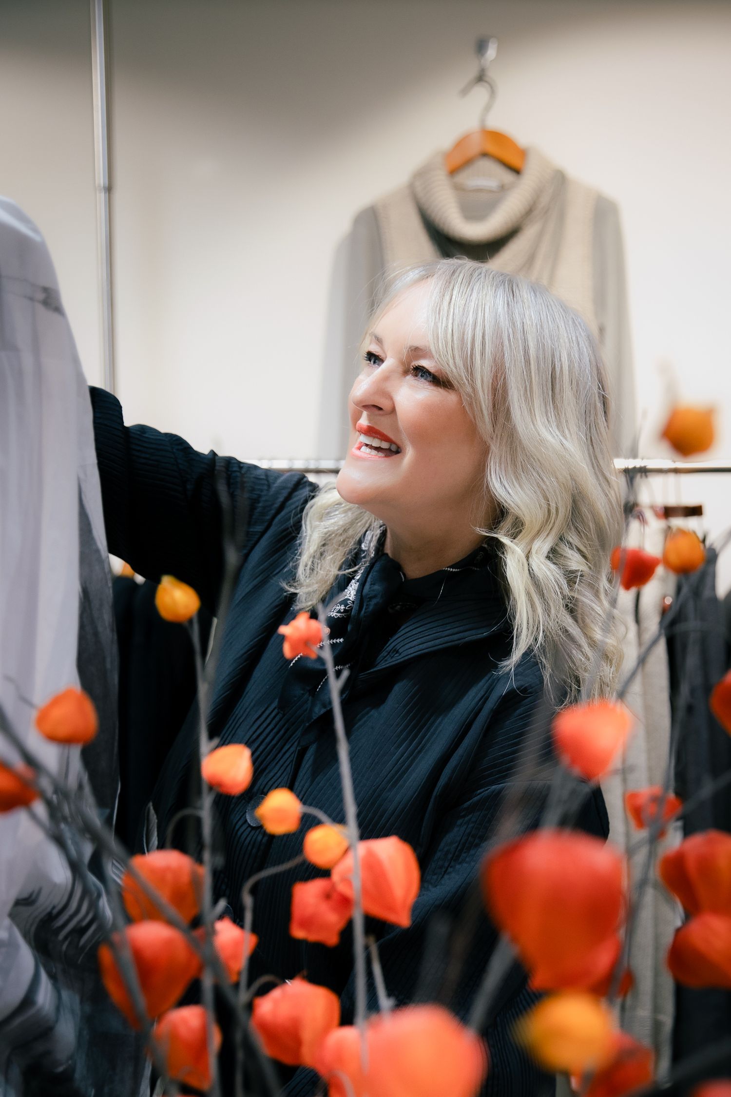 Smiling blonde woman browsing clothing rack surrounded by orange flowers in a boutique store.