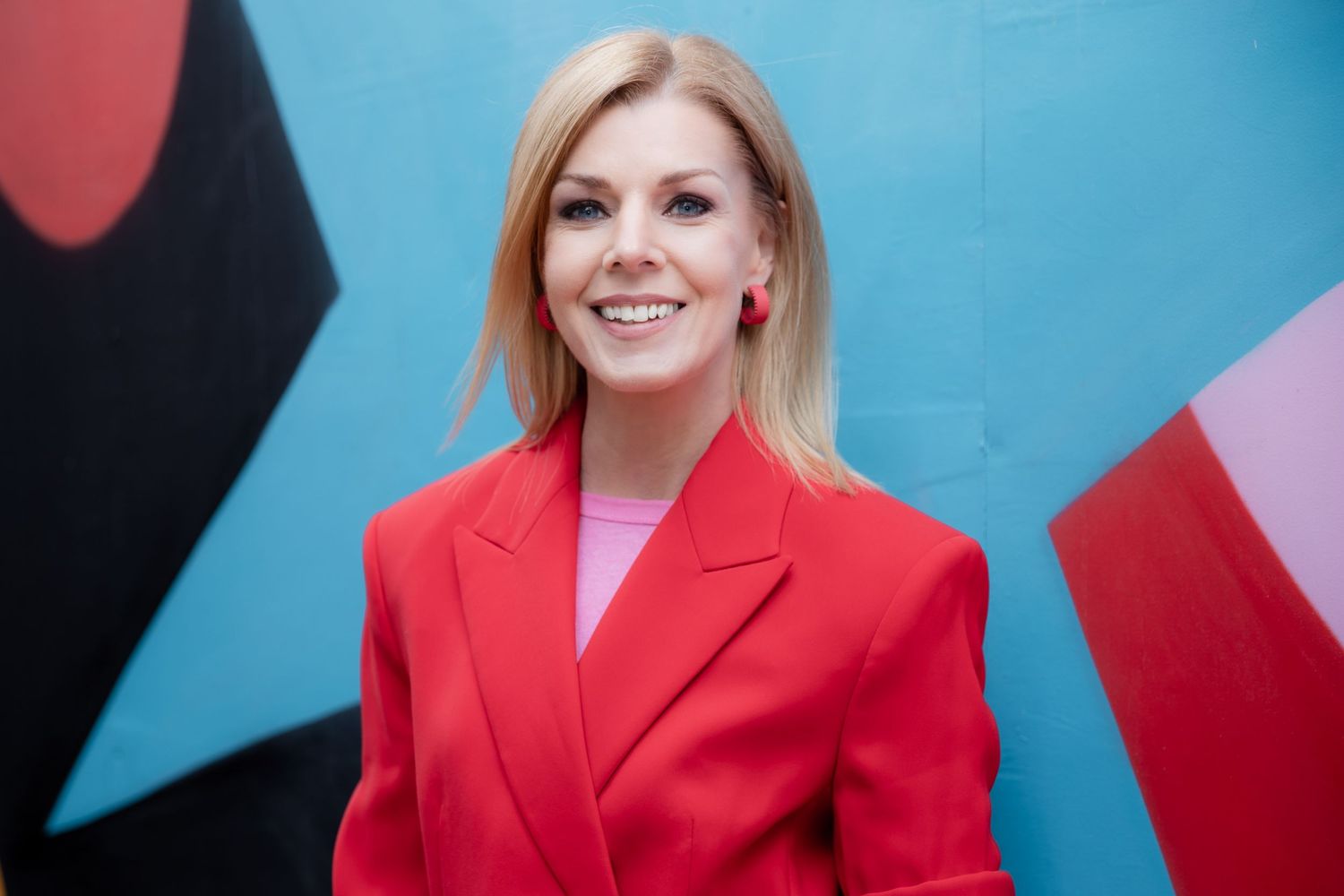 Smiling blonde woman in red blazer poses against colorful blue and red abstract mural background.