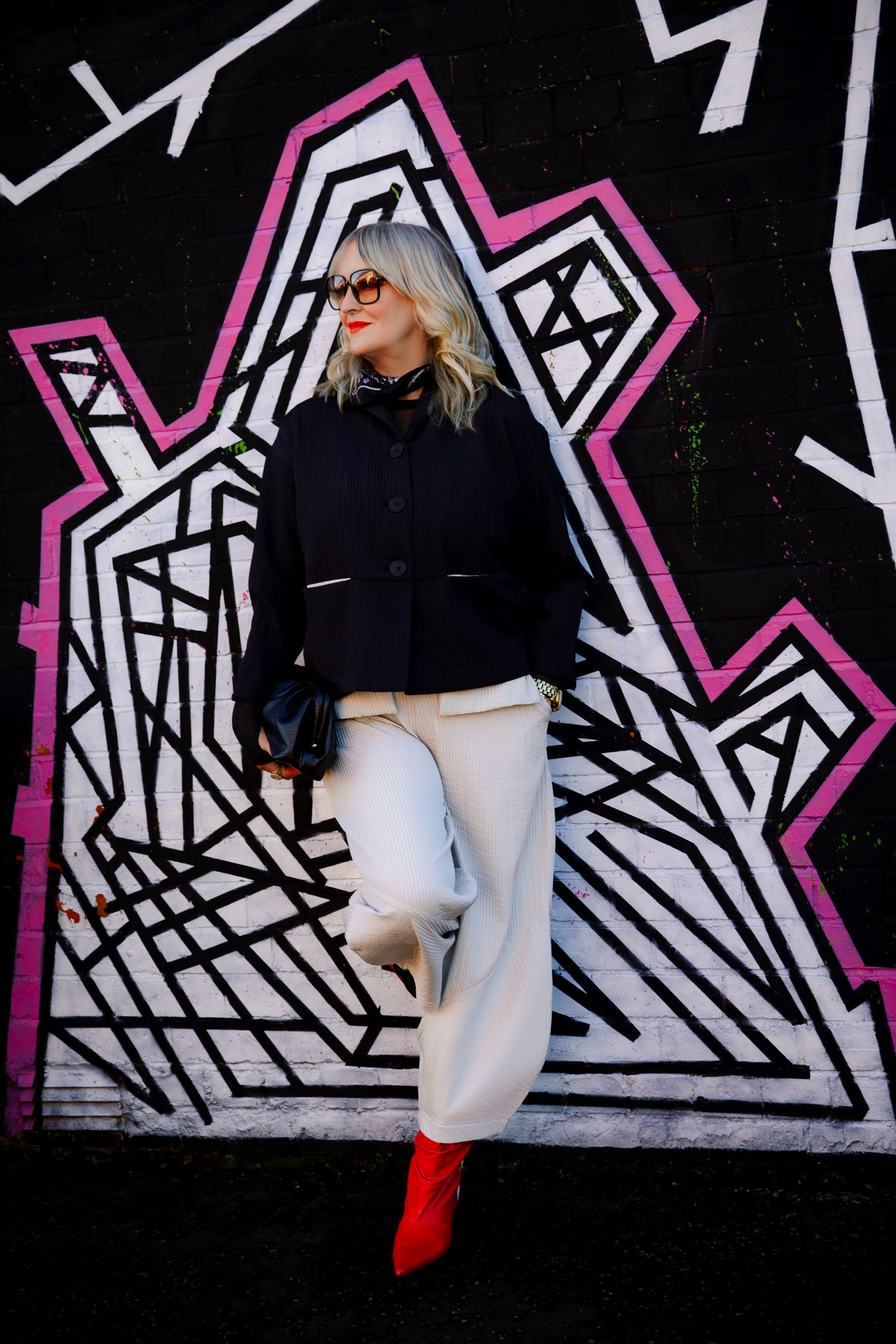 Woman in navy jacket, cream trousers, and red boots poses against bold black, white, and pink graffiti wall.