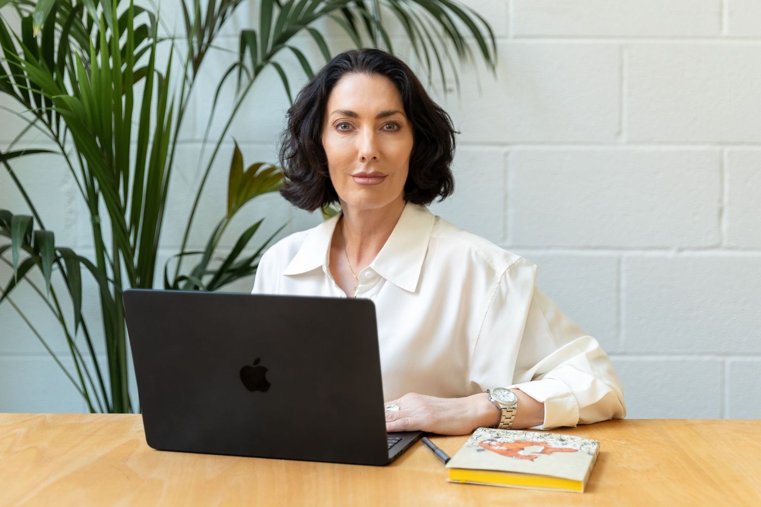 Woman in white blouse working on black MacBook at wooden desk with tropical plant background.