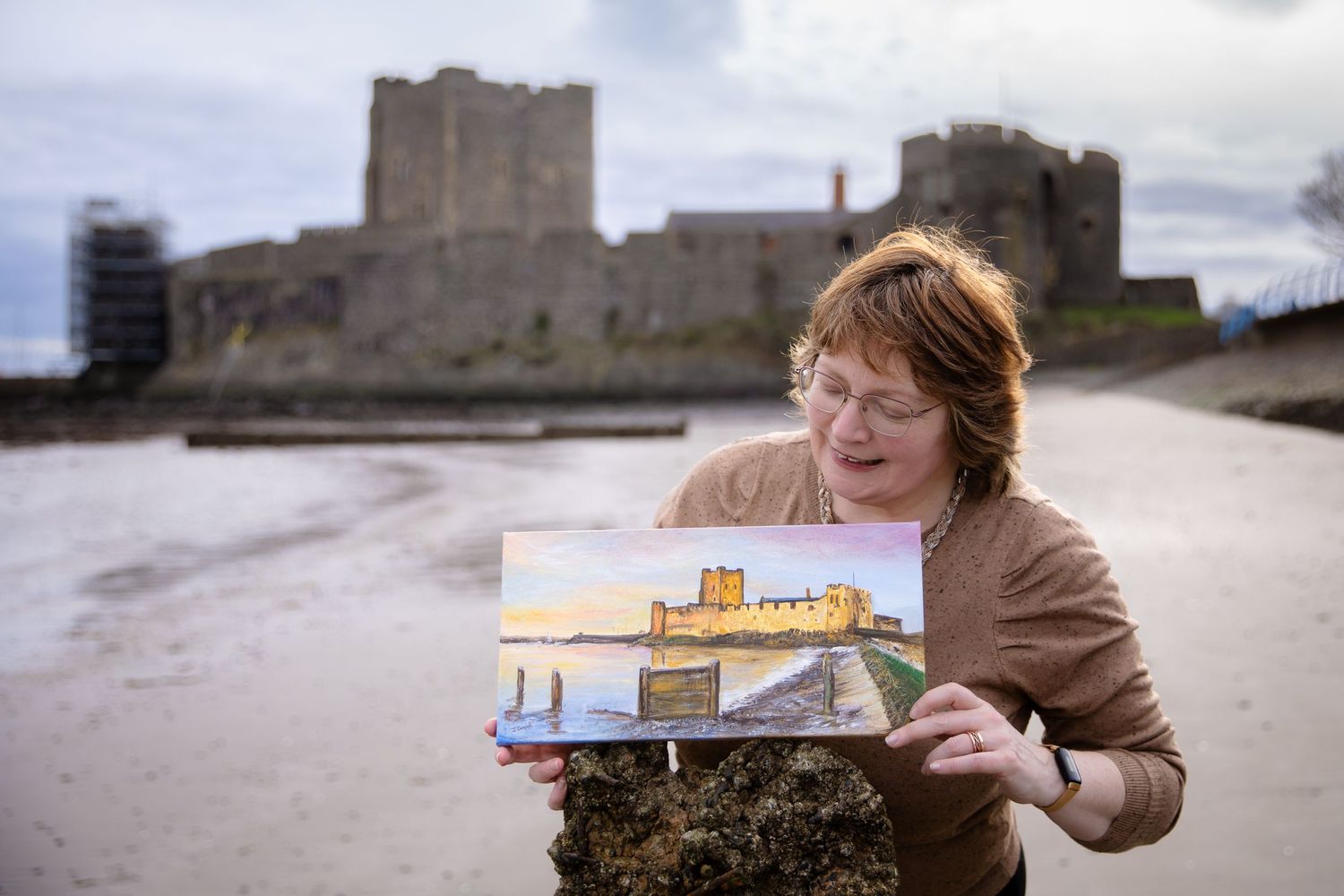 Woman holding a painting of Carrickfergus Castle on the beach with the historic castle in the background.