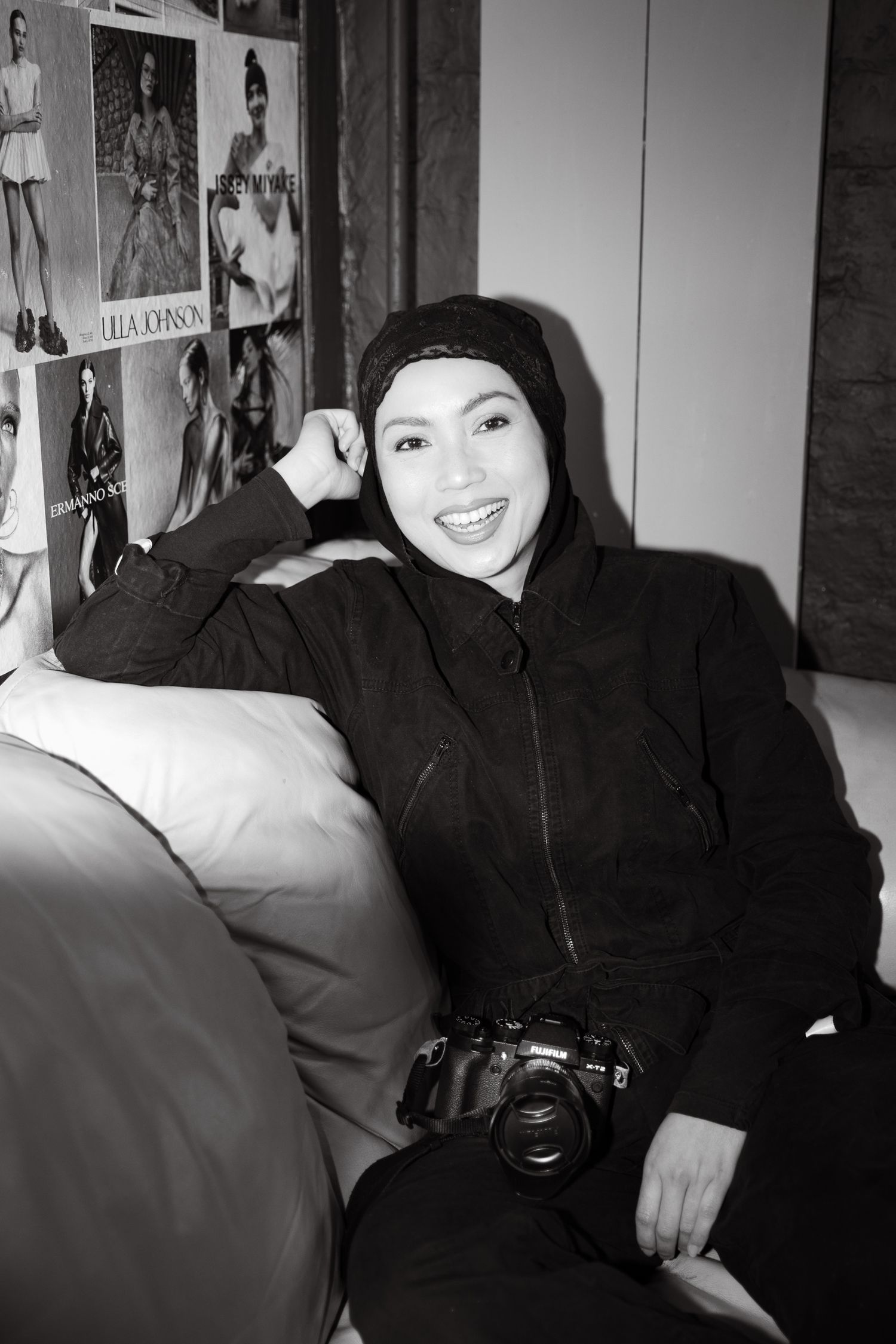 Black and white photo of a smiling woman in a hijab and black jacket holding a camera, sitting against a wall with fashion photos.