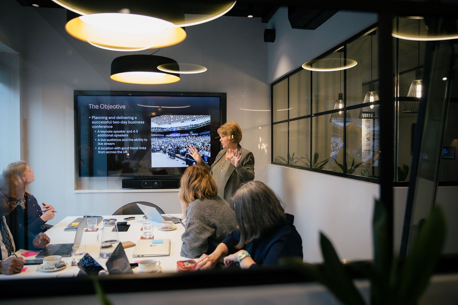 Business meeting in modern conference room with presenter at TV screen showing objectives to seated colleagues.