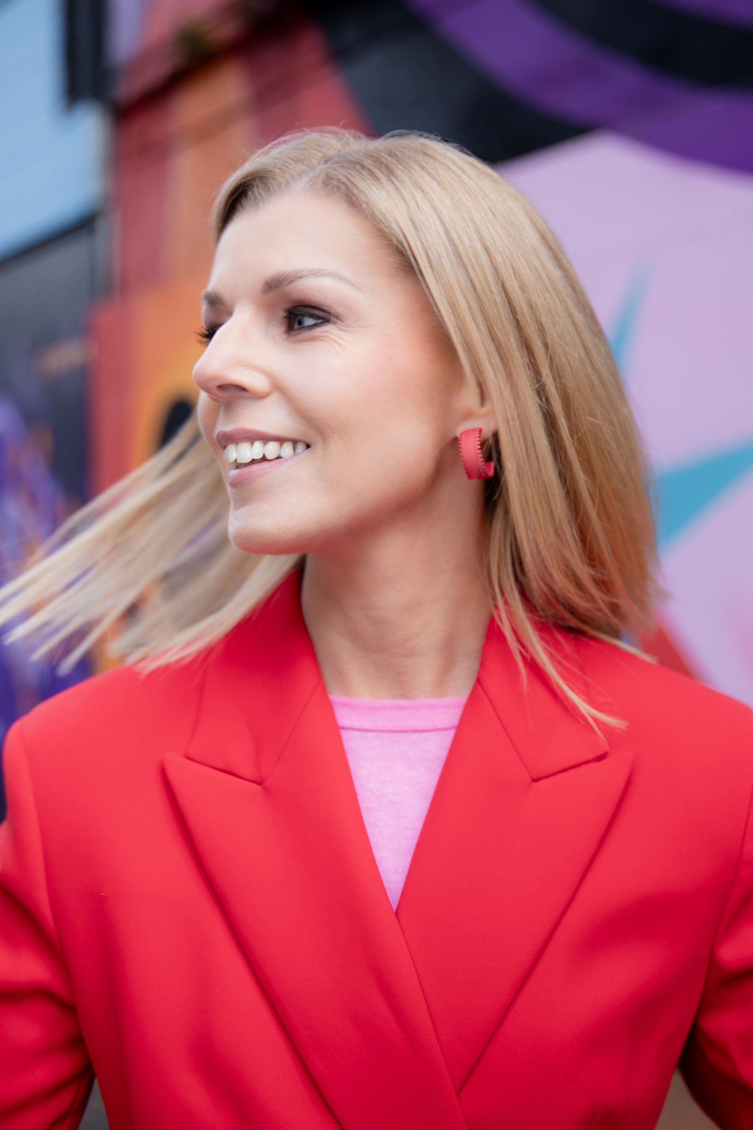 Smiling blonde woman in red blazer and pink top posing in front of colorful graffiti wall outdoors.