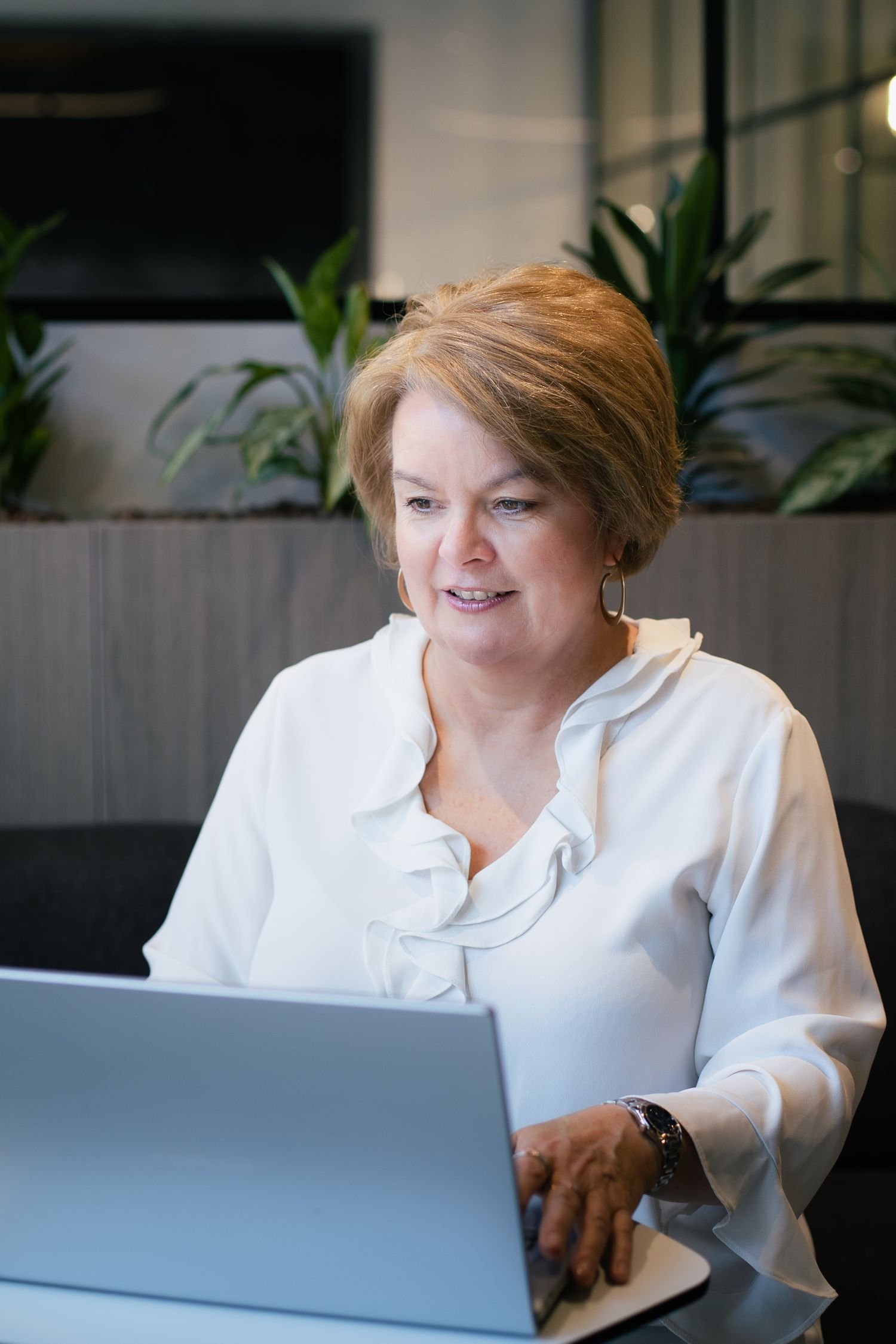 Professional woman in white blouse working on laptop in modern office with green plants.