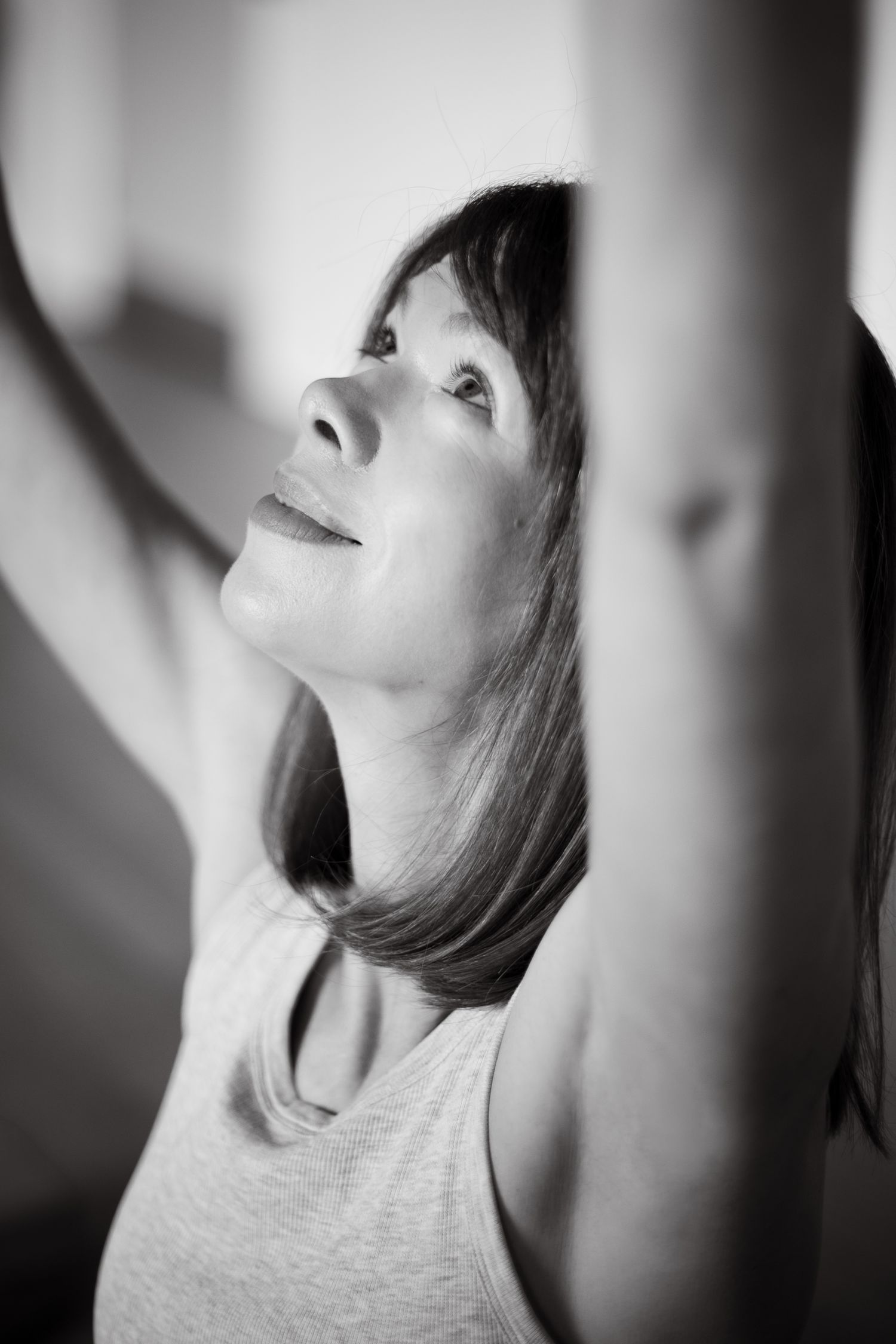 Black and white photo of a woman stretching arms overhead, looking up with a serene expression in a yoga pose.