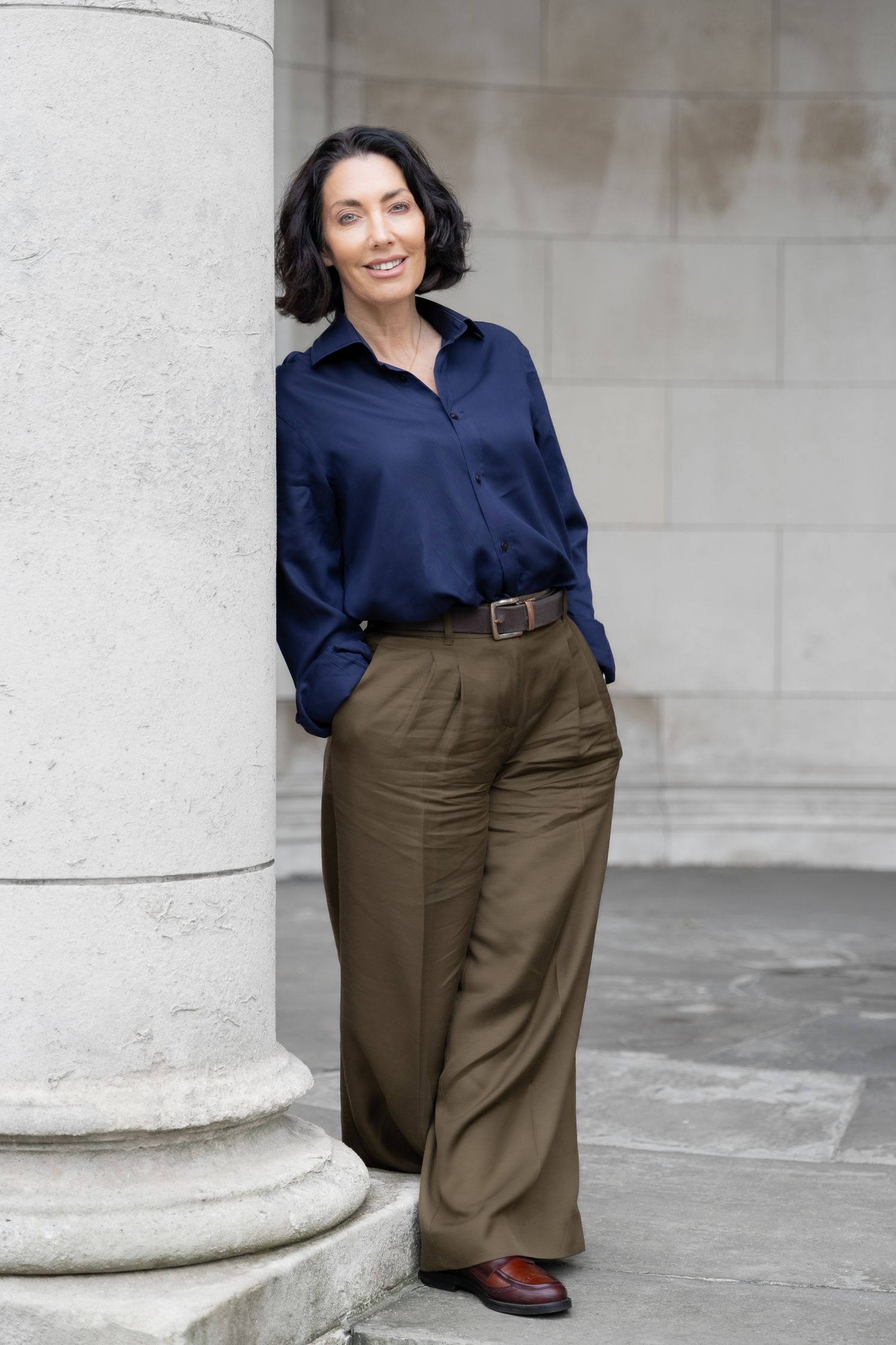 Woman in navy blouse and olive wide-leg trousers leaning against a stone column outdoors.