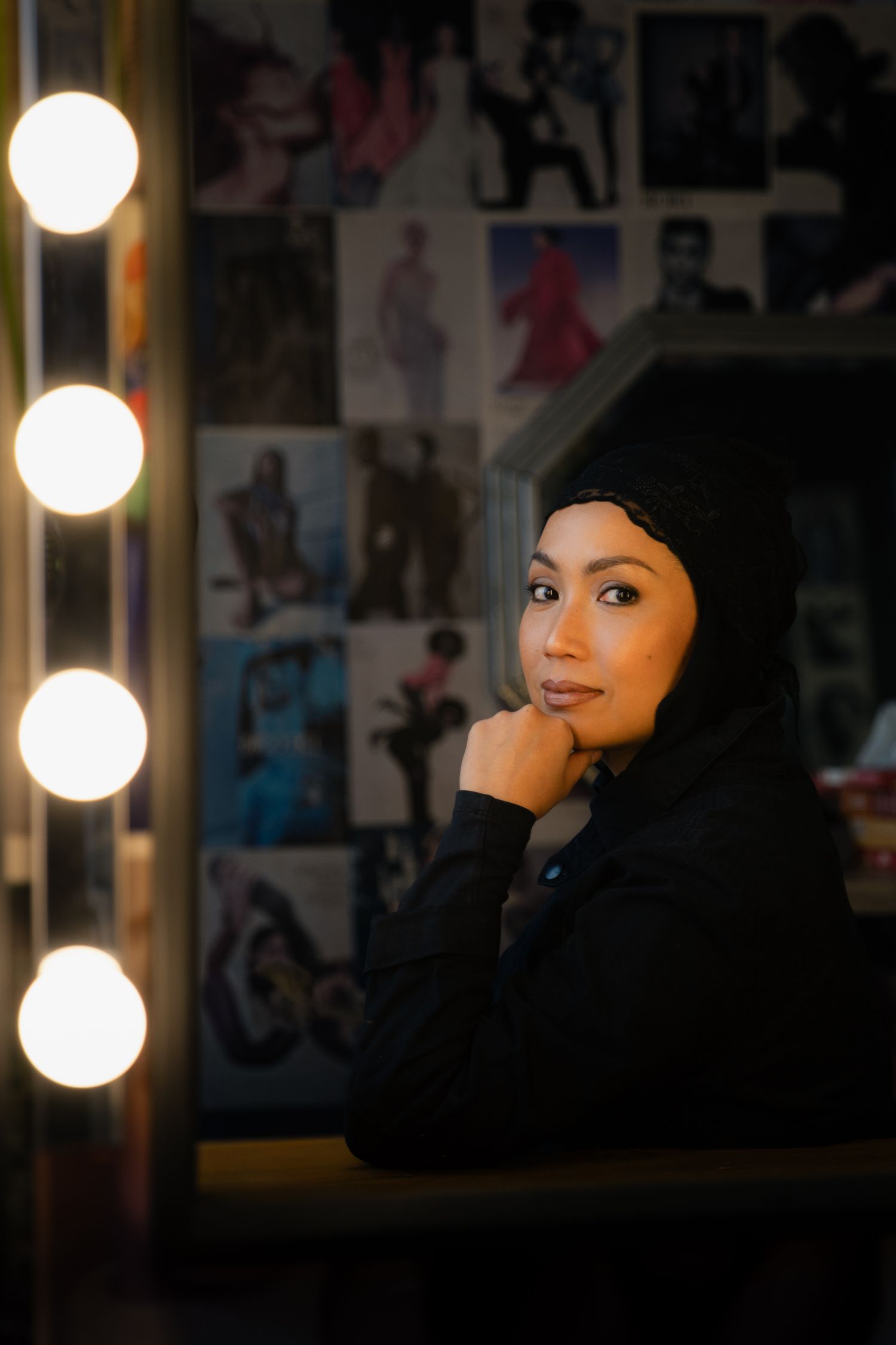 Woman in black hijab poses thoughtfully before a vanity mirror with lights, surrounded by colorful posters.