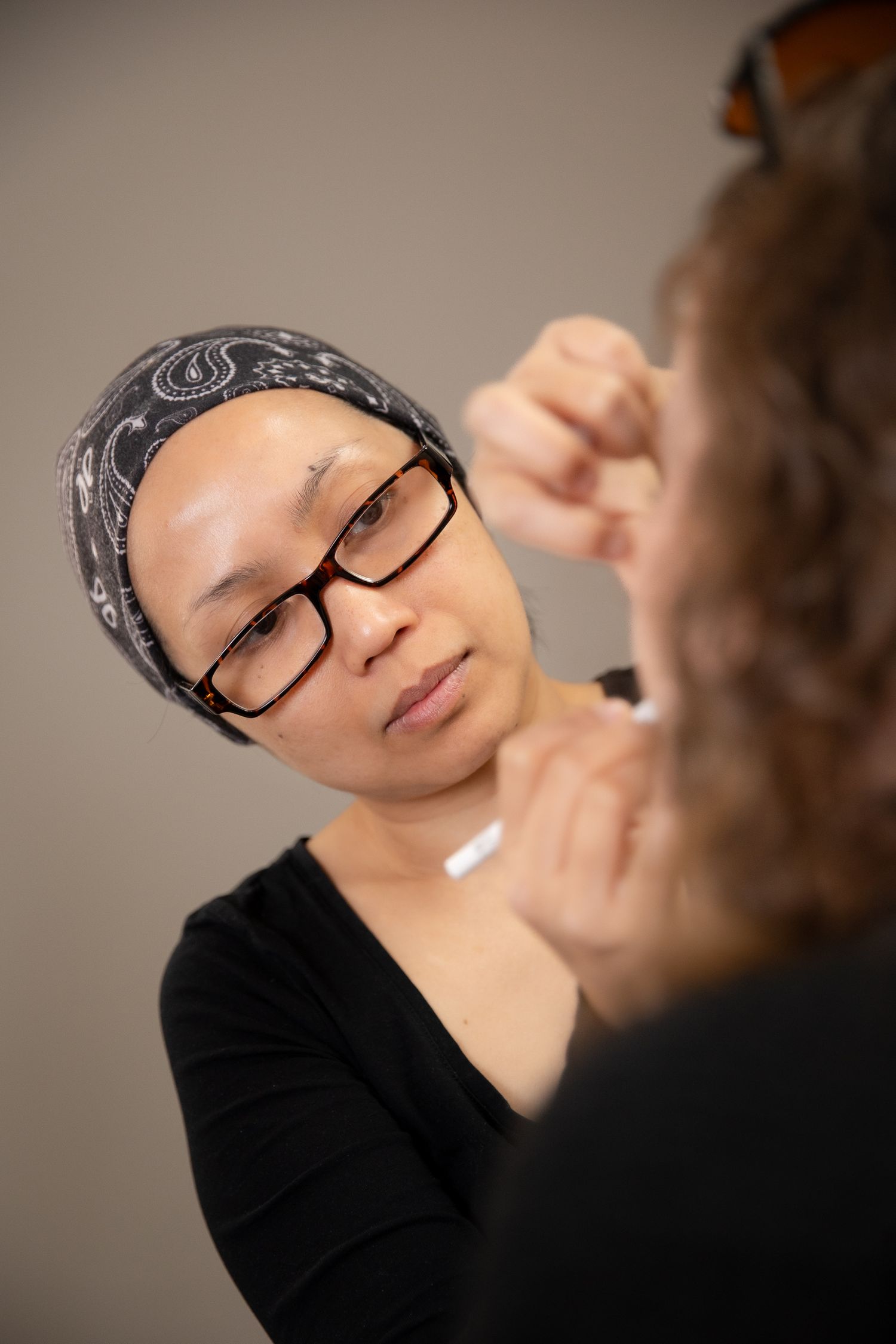 A makeup artist wearing glasses and a bandana applies makeup to a client in a professional setting.