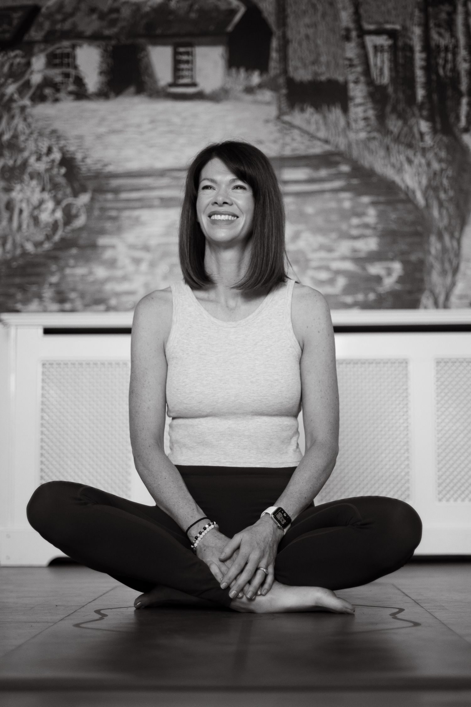Smiling woman sitting cross-legged on yoga mat in black and white photo, wearing tank top and leggings.