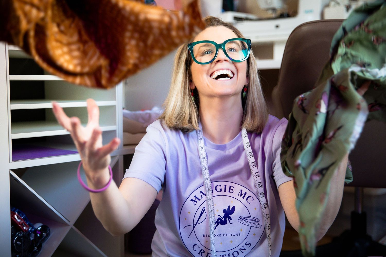 Woman in teal glasses and Margie MC t-shirt laughing joyfully while tossing fabric in a creative studio.