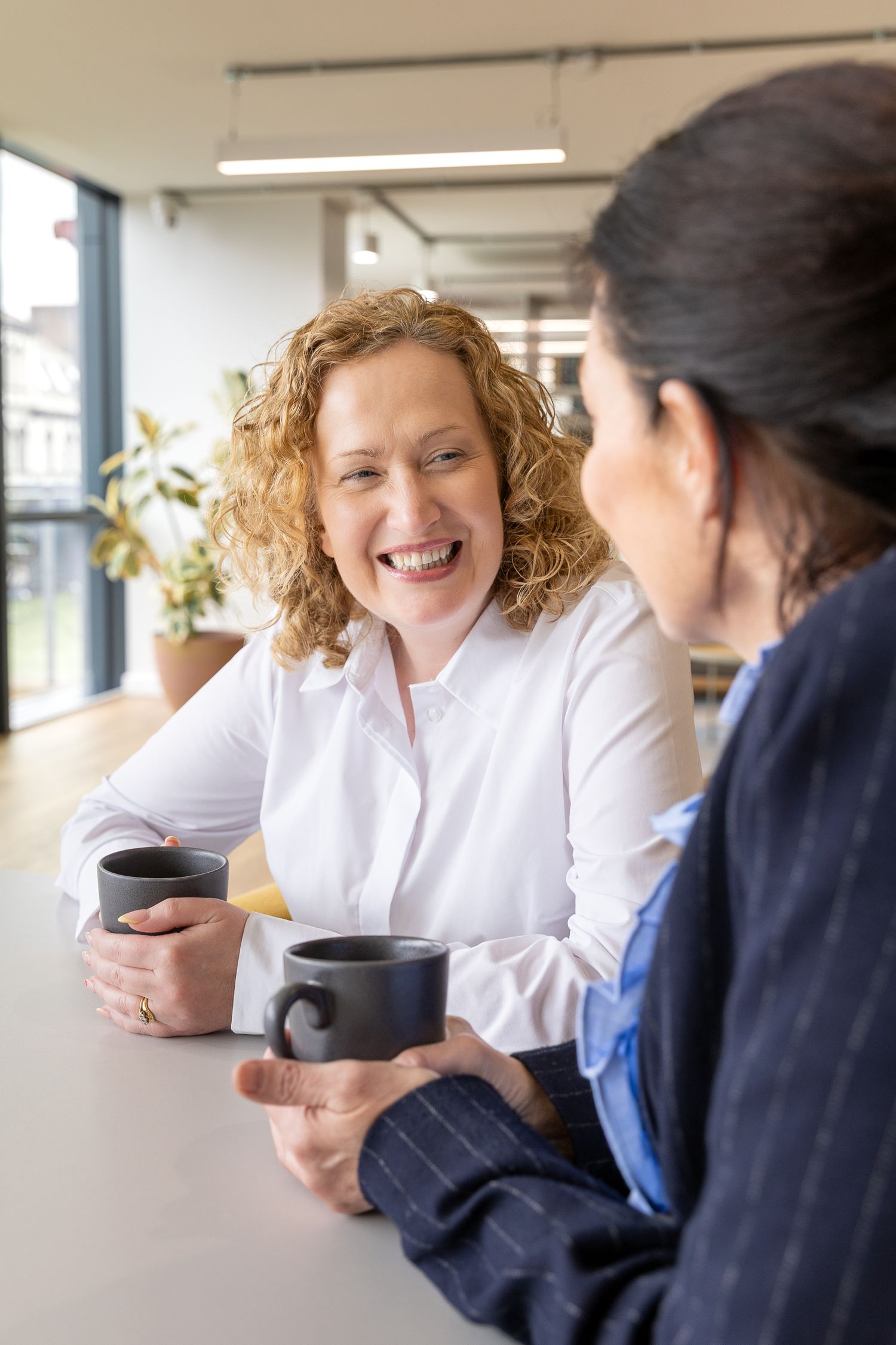 Two professional women laughing and sharing coffee mugs in a bright modern office setting.