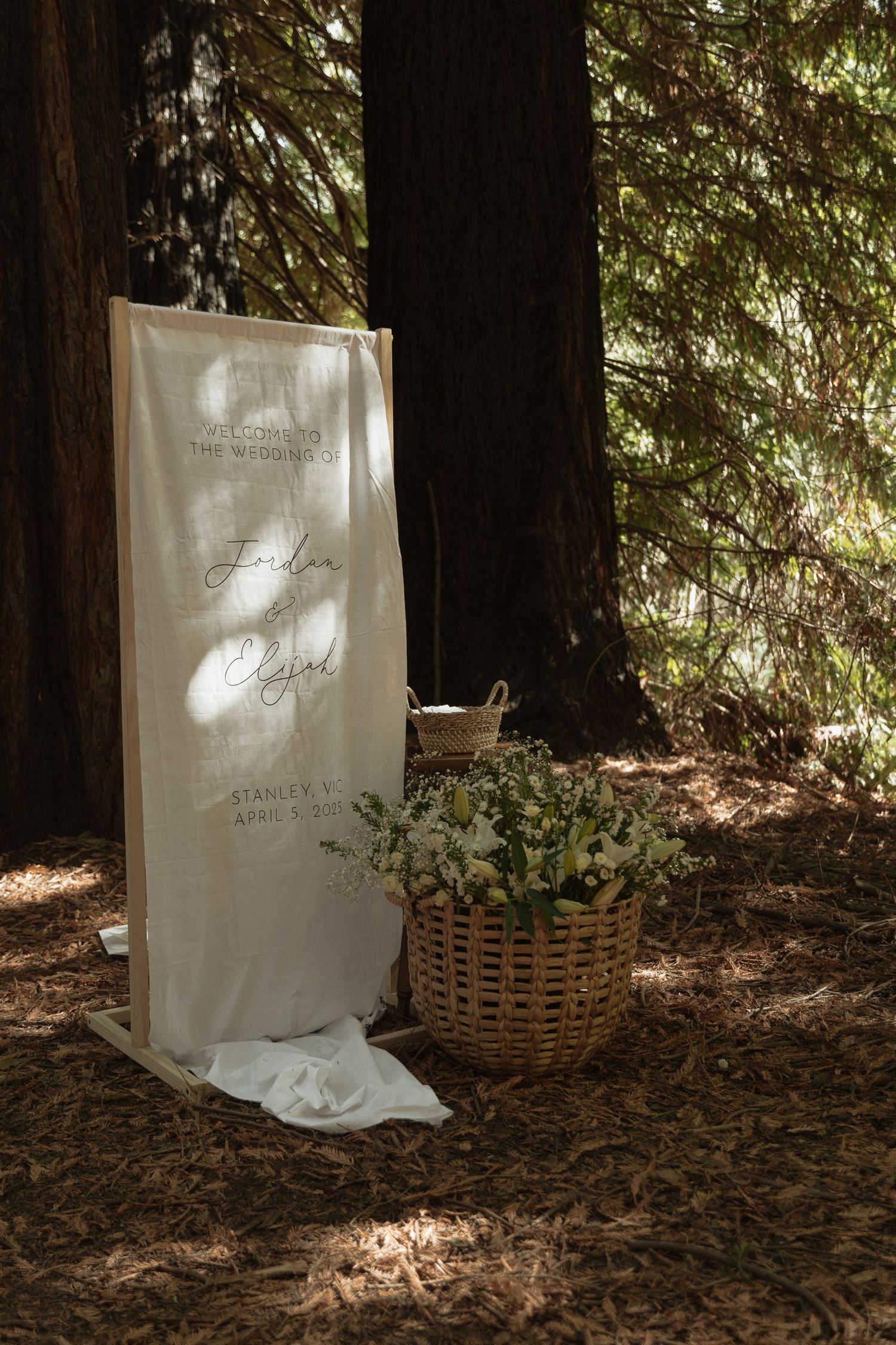 A white fabric banner and wicker basket with flowers sit in dappled sunlight on a forest floor covered in pine needles.