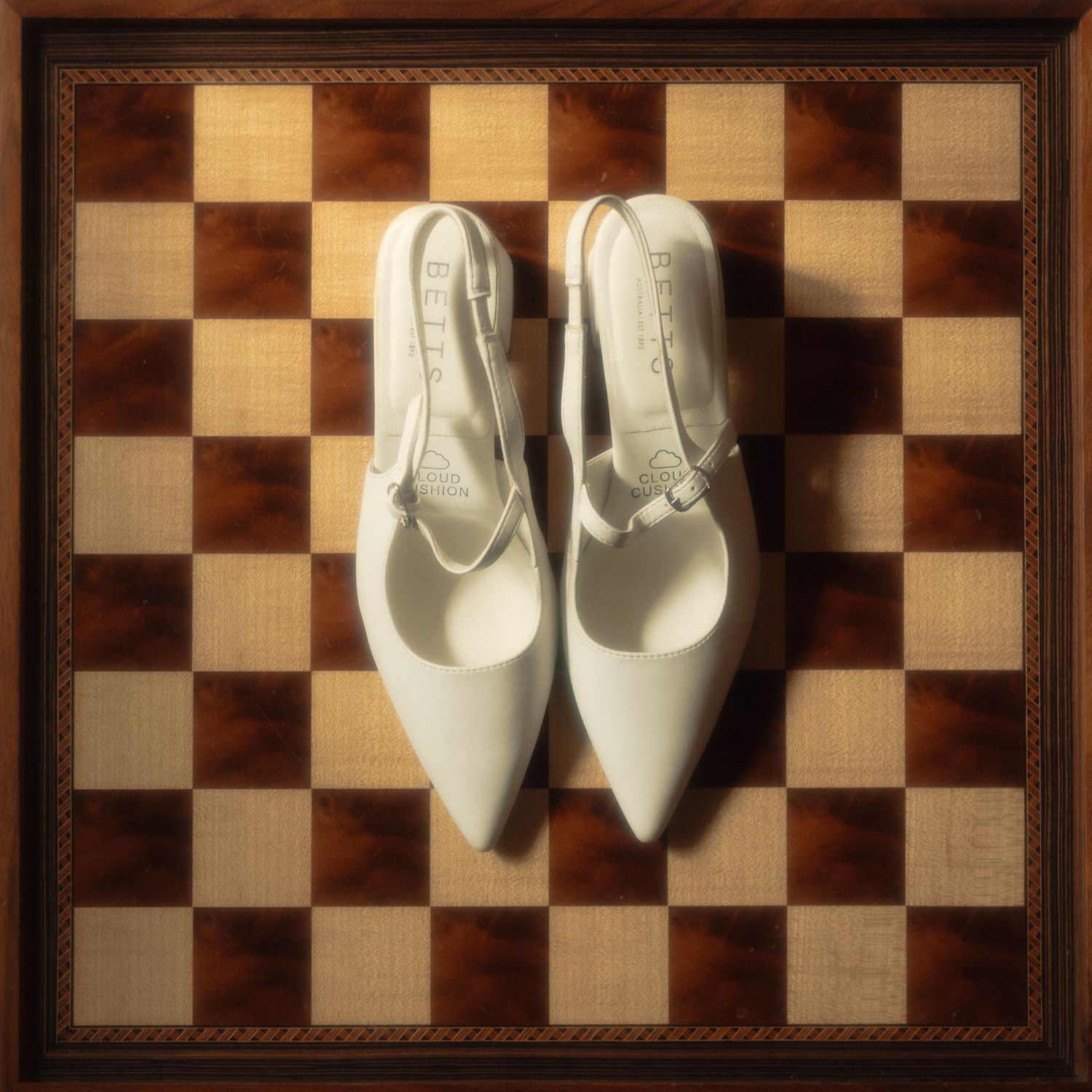 A pair of elegant white slingback heels placed on a brown and beige checkered chess board pattern surface.