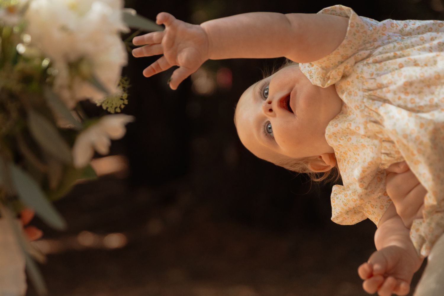 A small child wearing a lacy white dress reaches upward with an outstretched hand in warm lighting.