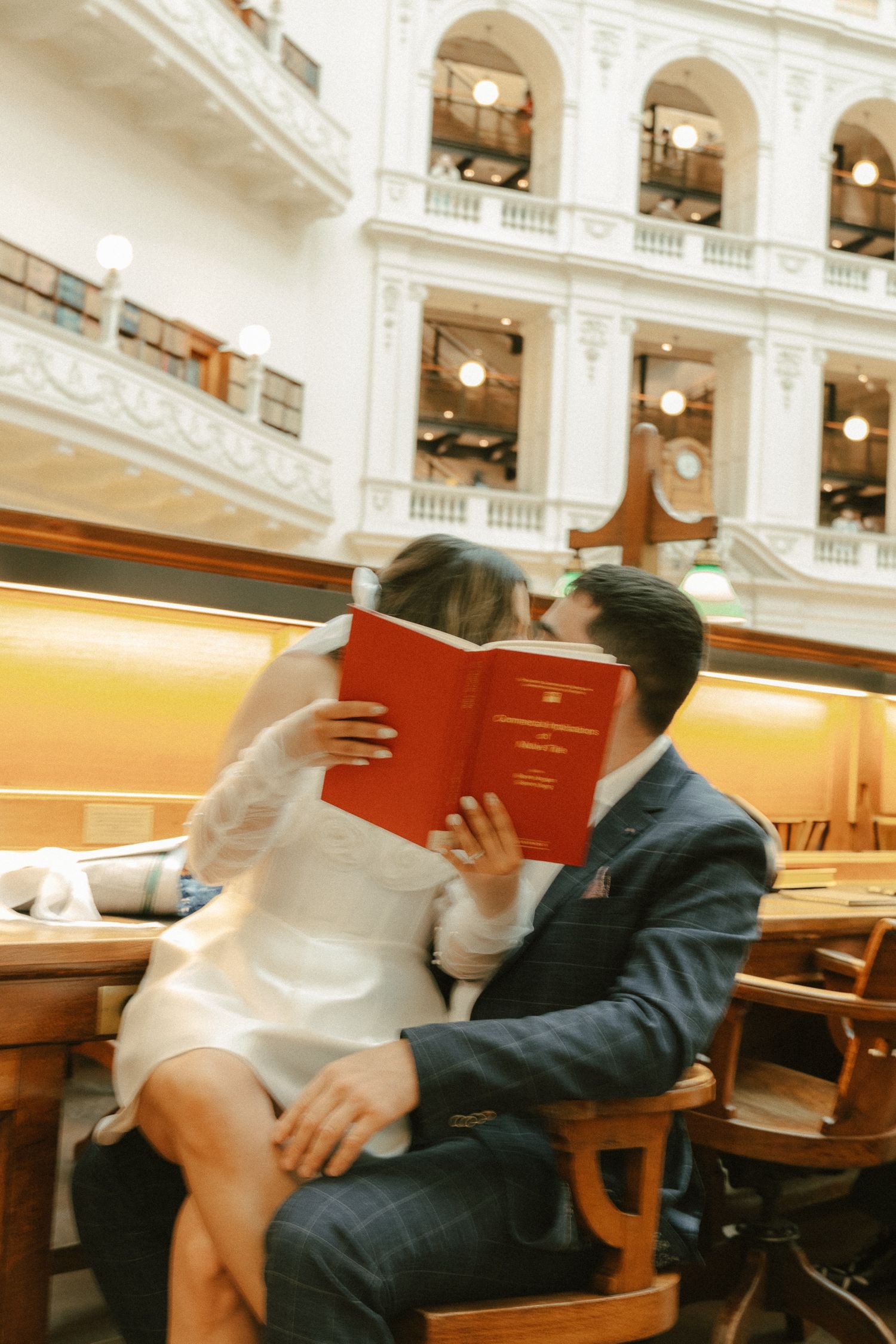 A couple shares a romantic moment behind a red book in an elegant vintage building with arched architecture.