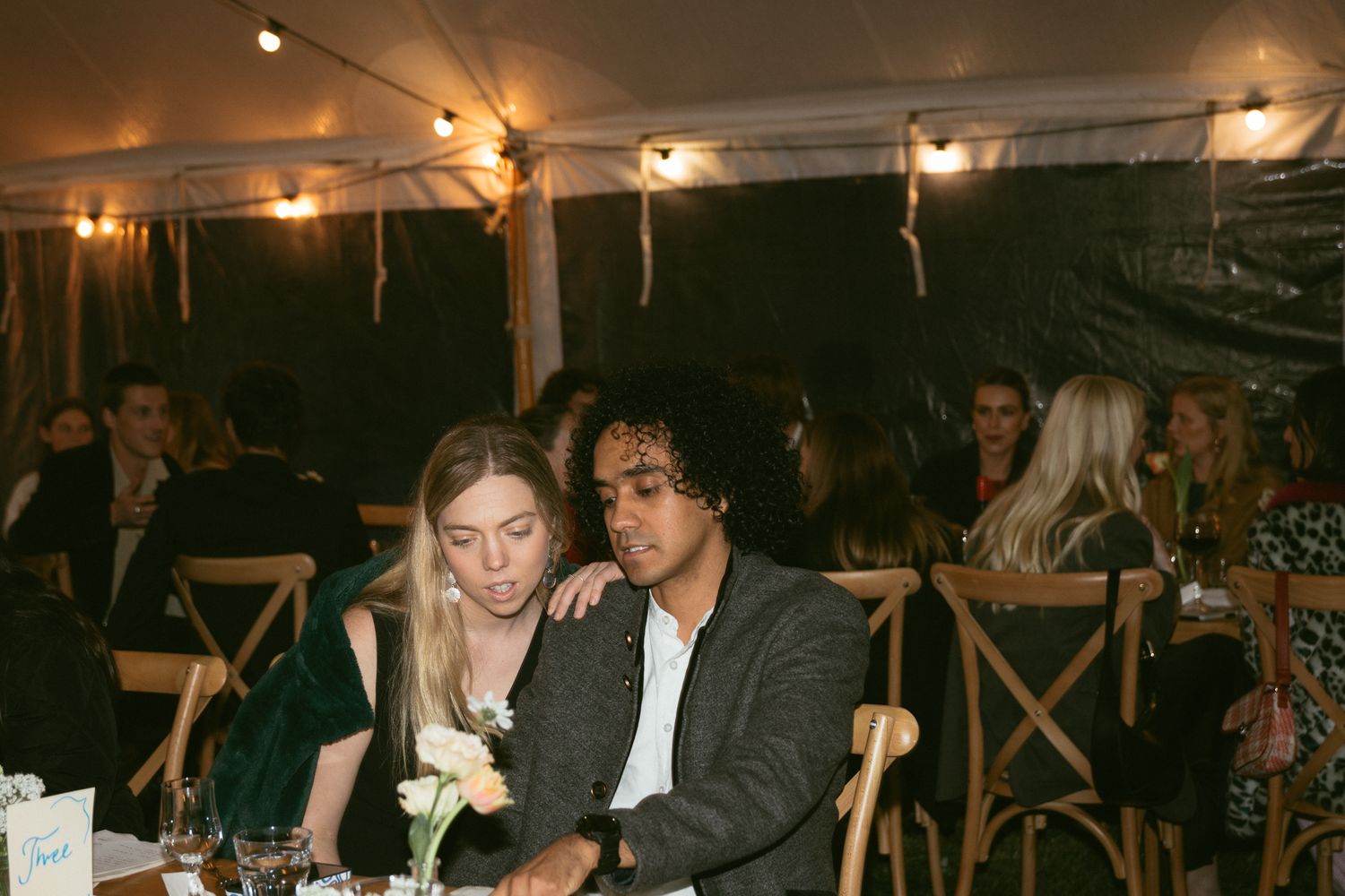 A couple sits together at a rustic outdoor reception with string lights overhead and wooden chairs in the background.