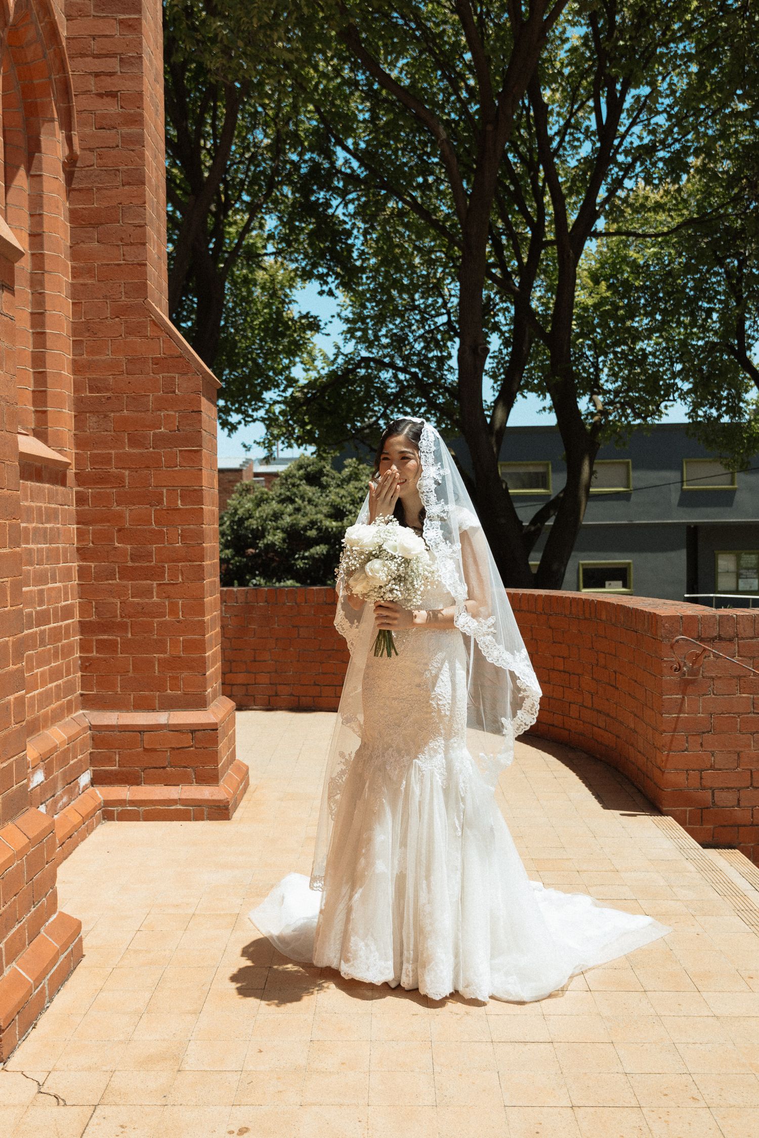 Bride in lace wedding gown and veil holds bouquet outside brick church on sunny day.