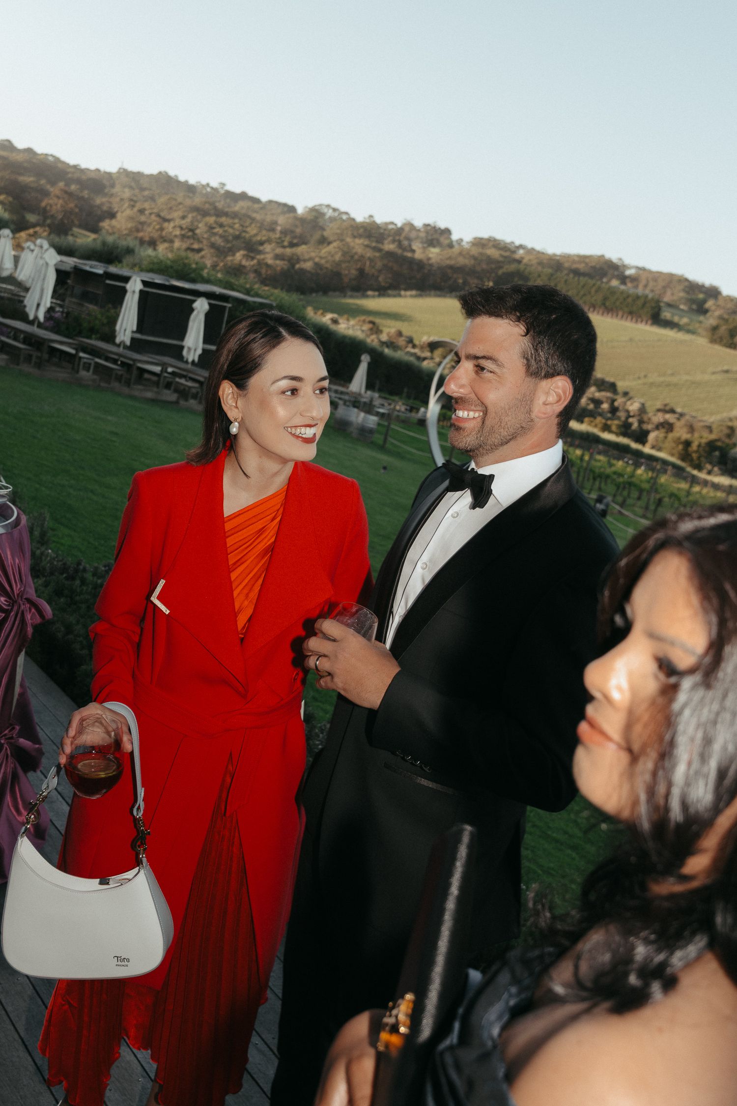 Couple in formal attire sharing a candid moment at an outdoor event with a red dress standing out against green scenery.