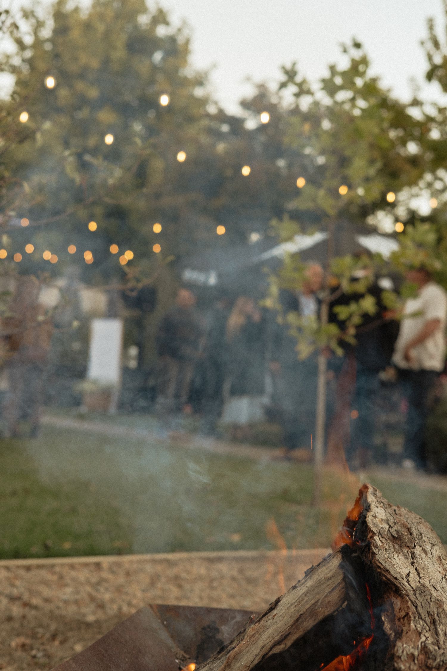 Burning campfire with glowing string lights and trees blurred in background creating cozy outdoor ambiance.
