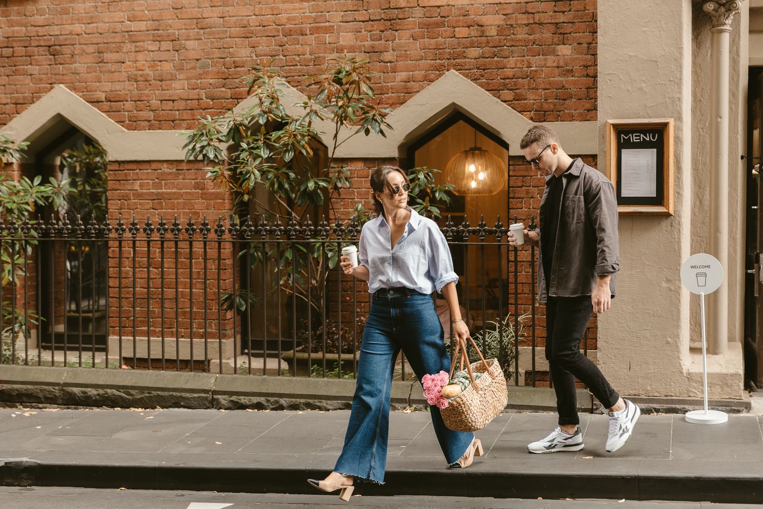 Two people walking down a city sidewalk past a brick building with an arched doorway and iron fence.