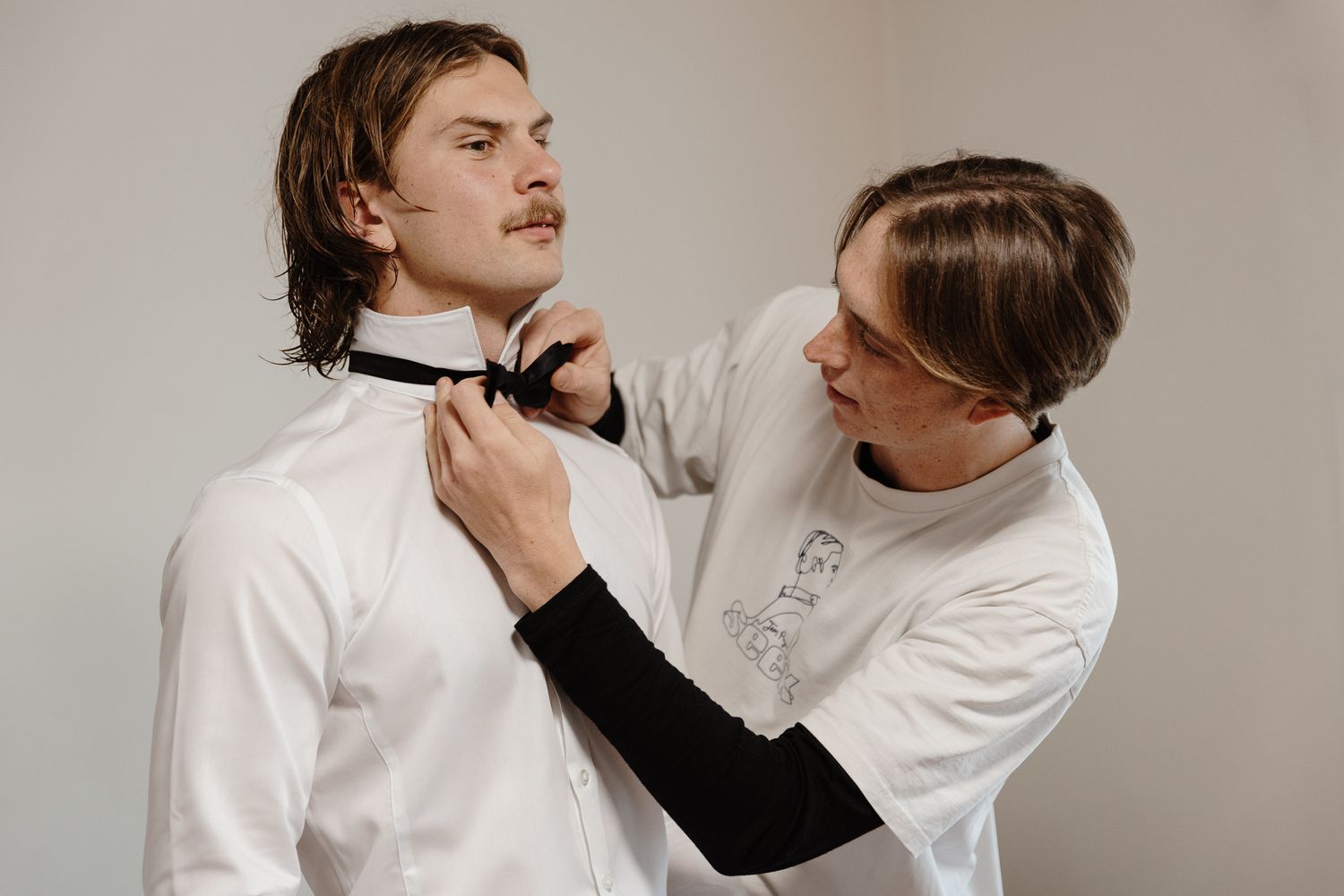 A person adjusts another person's bowtie while both wear white dress shirts in a formal styling session.