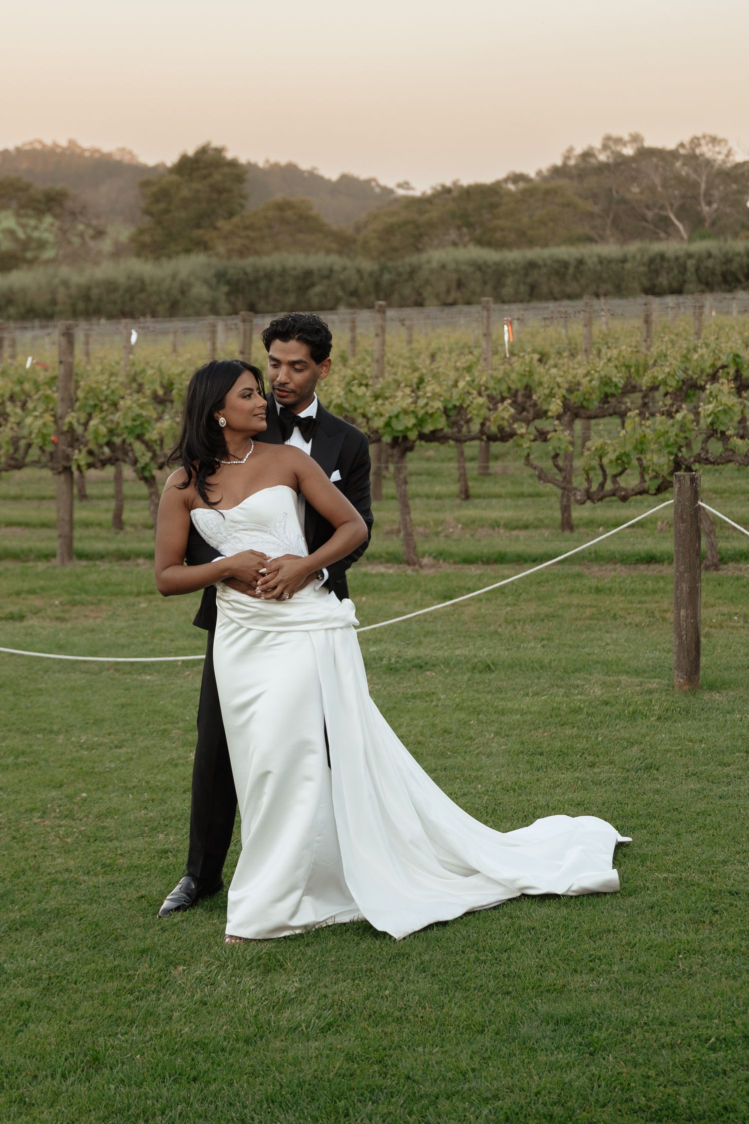 A couple shares romantic moments in a vineyard as they pose for wedding photos on a grassy field at sunset.