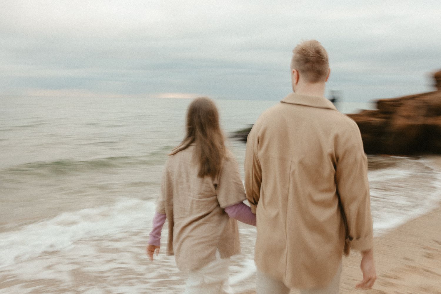 A couple walks along a misty beach shoreline in matching beige outfits.