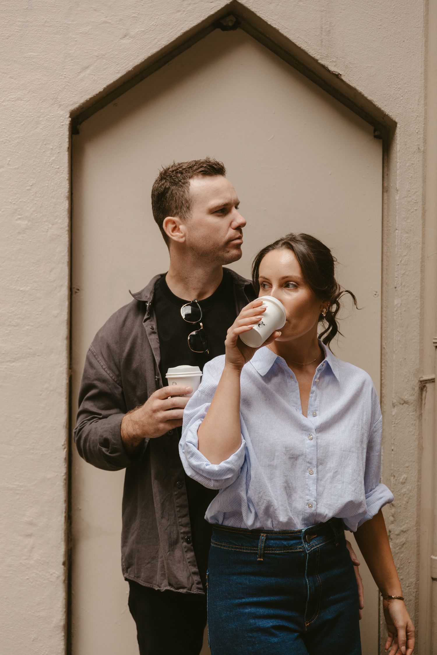 A couple stands drinking coffee together against a neutral wall with an architectural house-shaped detail behind them.