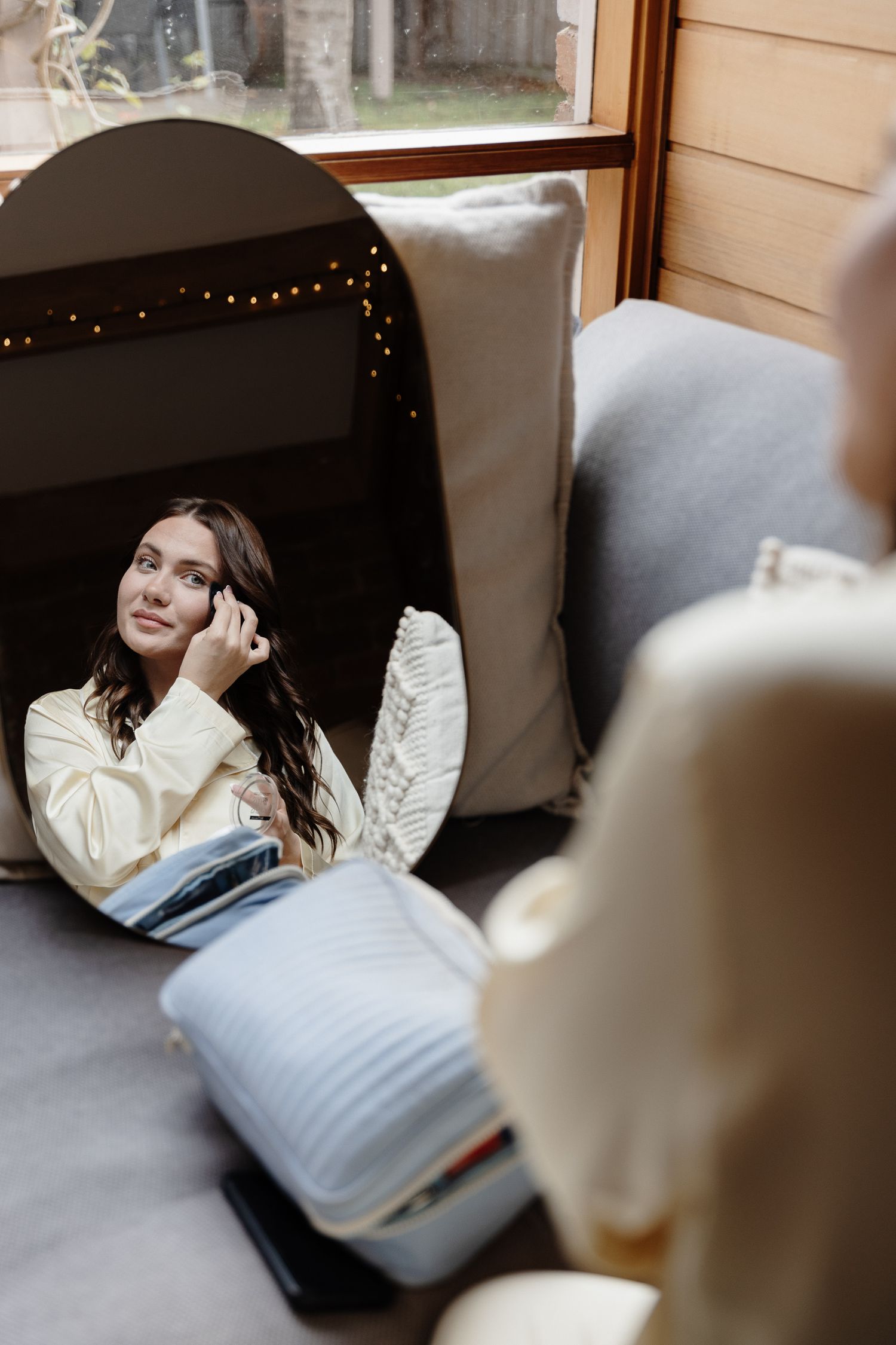 A reflection in an oval mirror showing a window seat with throw pillows and fairy lights in the background.