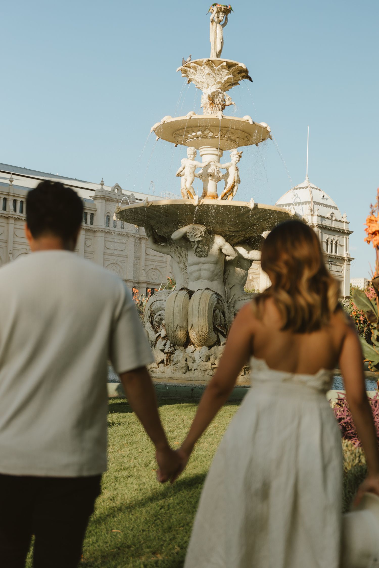 A couple admires an ornate classical fountain in a garden setting with white buildings in the background.