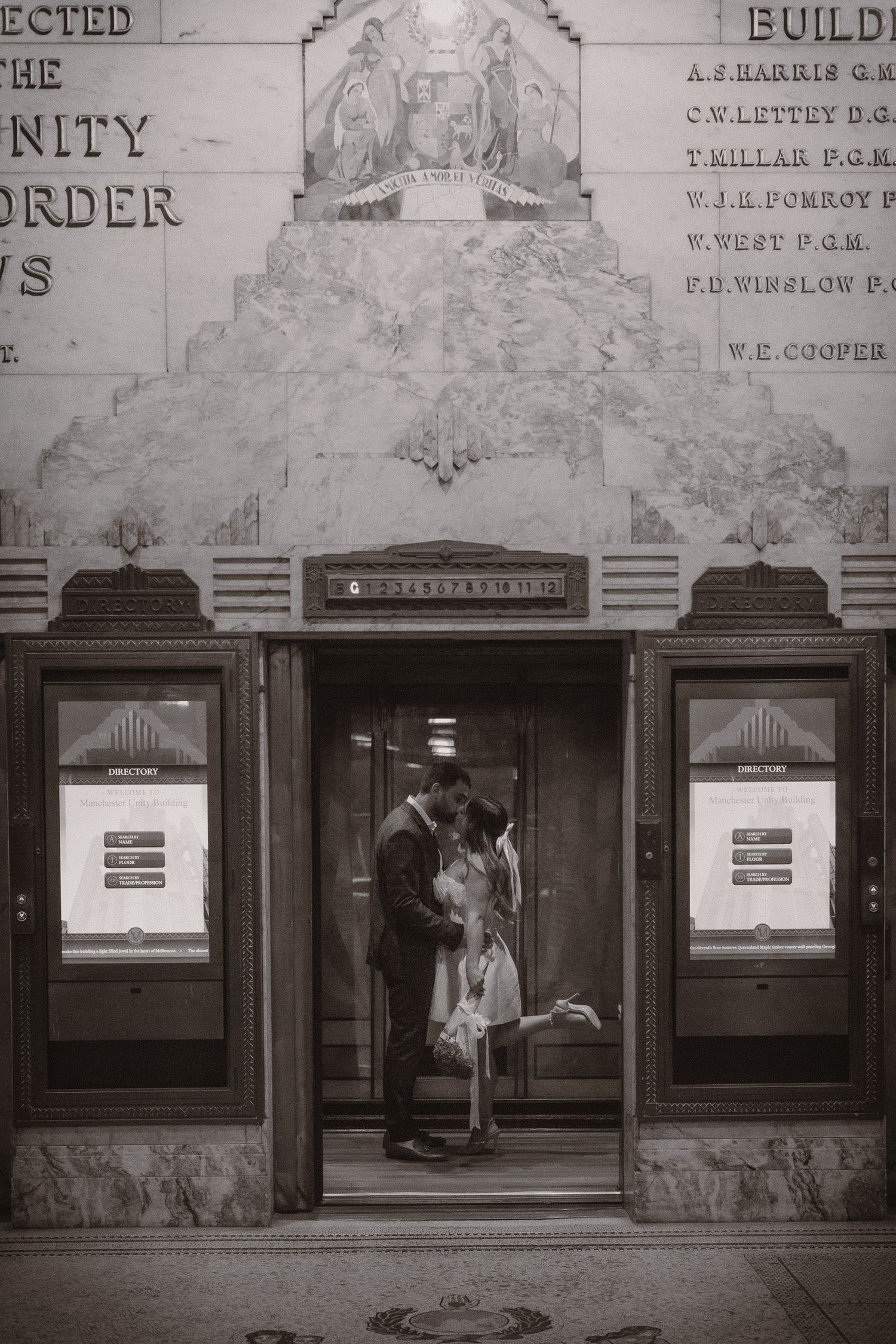 Two figures stand in the doorway entrance of a vintage building with old fashioned ticket windows on either side.