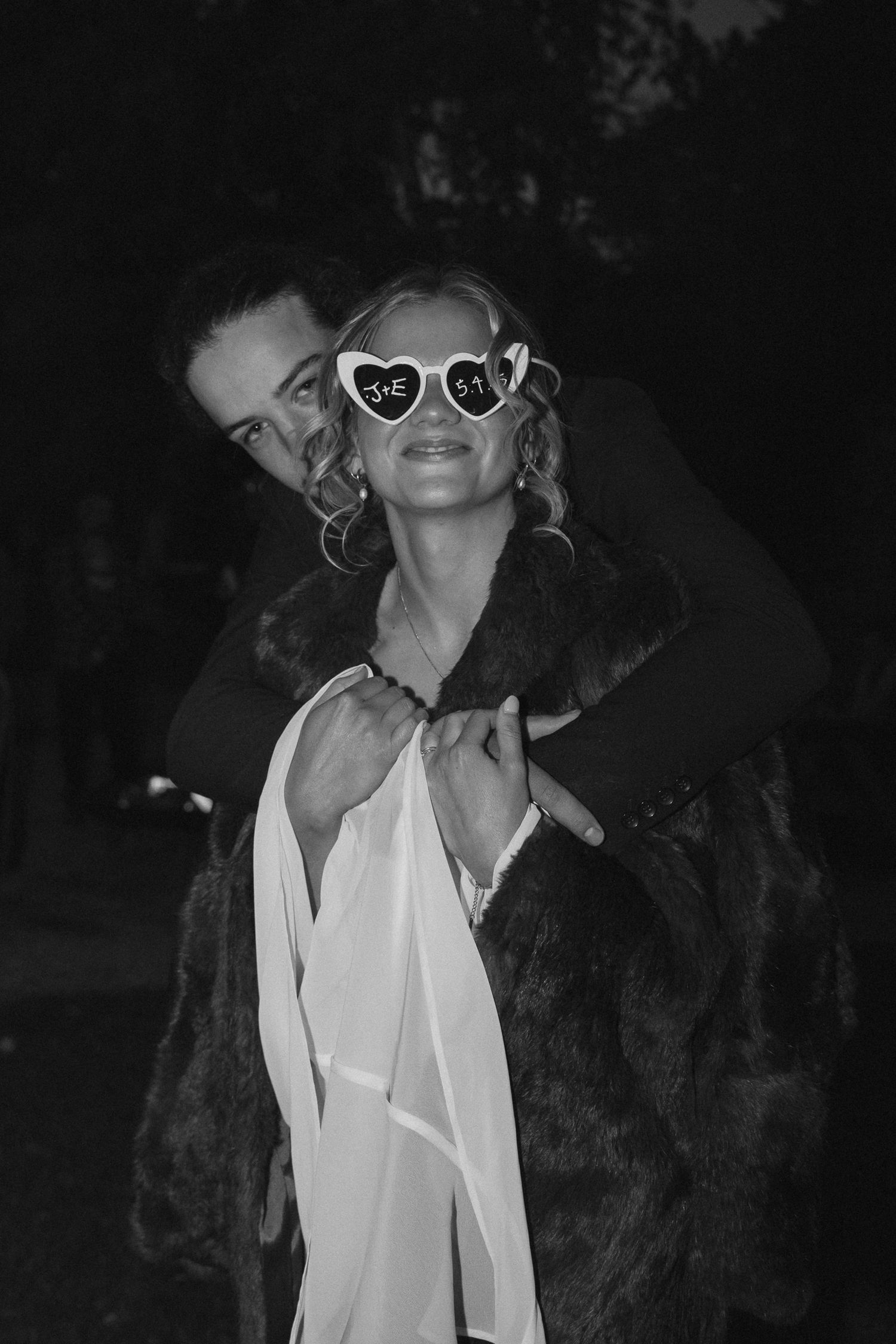 Black and white photo of a figure in heart-shaped sunglasses and fur coat being embraced from behind at night.