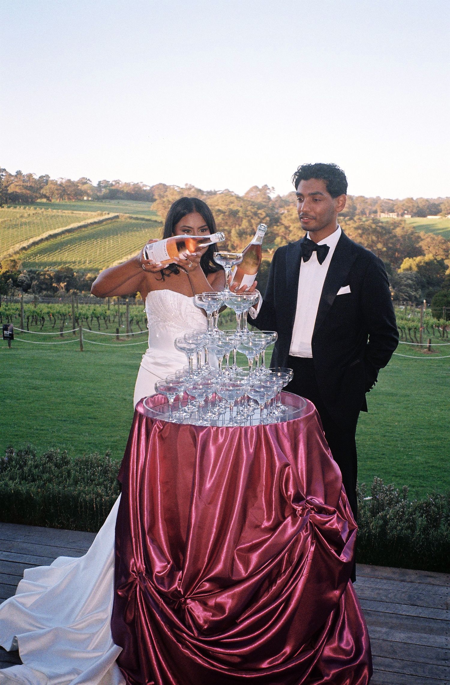 Champagne tower setup with burgundy satin draping on a table overlooking vineyard views at sunset.