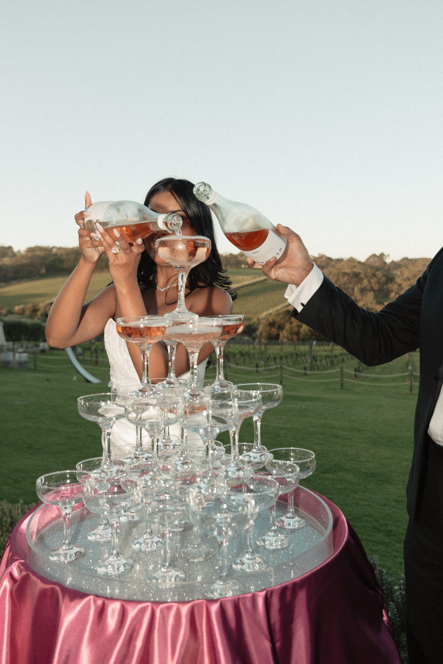 Wedding party pours champagne into tower of crystal glasses stacked on pink tablecloth outdoors.