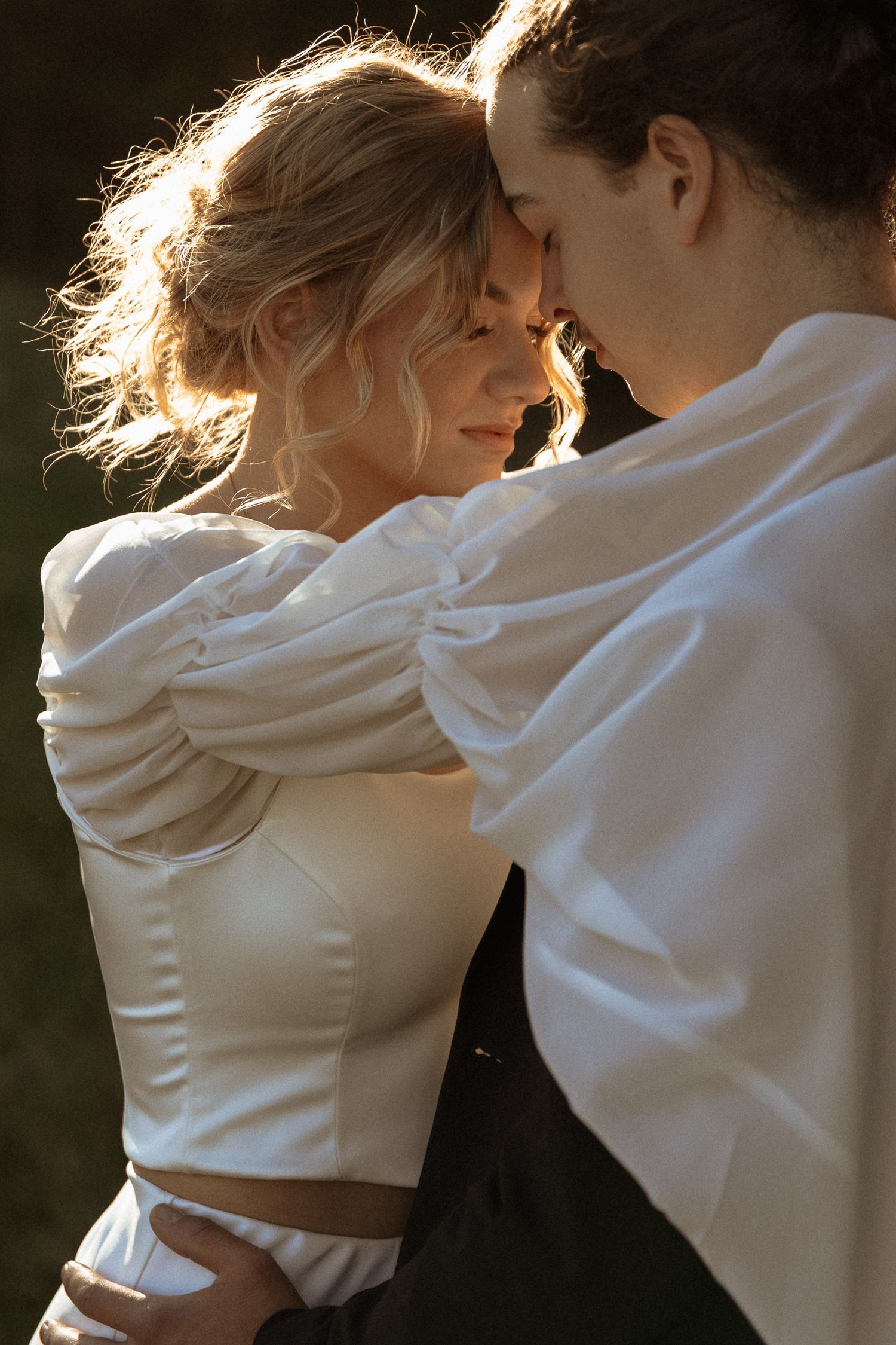 Romantic golden hour portrait of a couple in white clothing sharing an intimate moment against sunlit background.