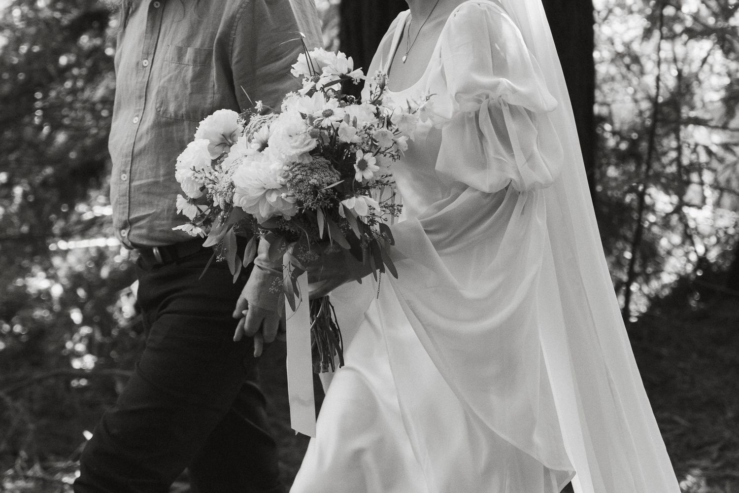 Black and white wedding photo showing dress details and floral bouquet with flowing white fabric and ribbons.