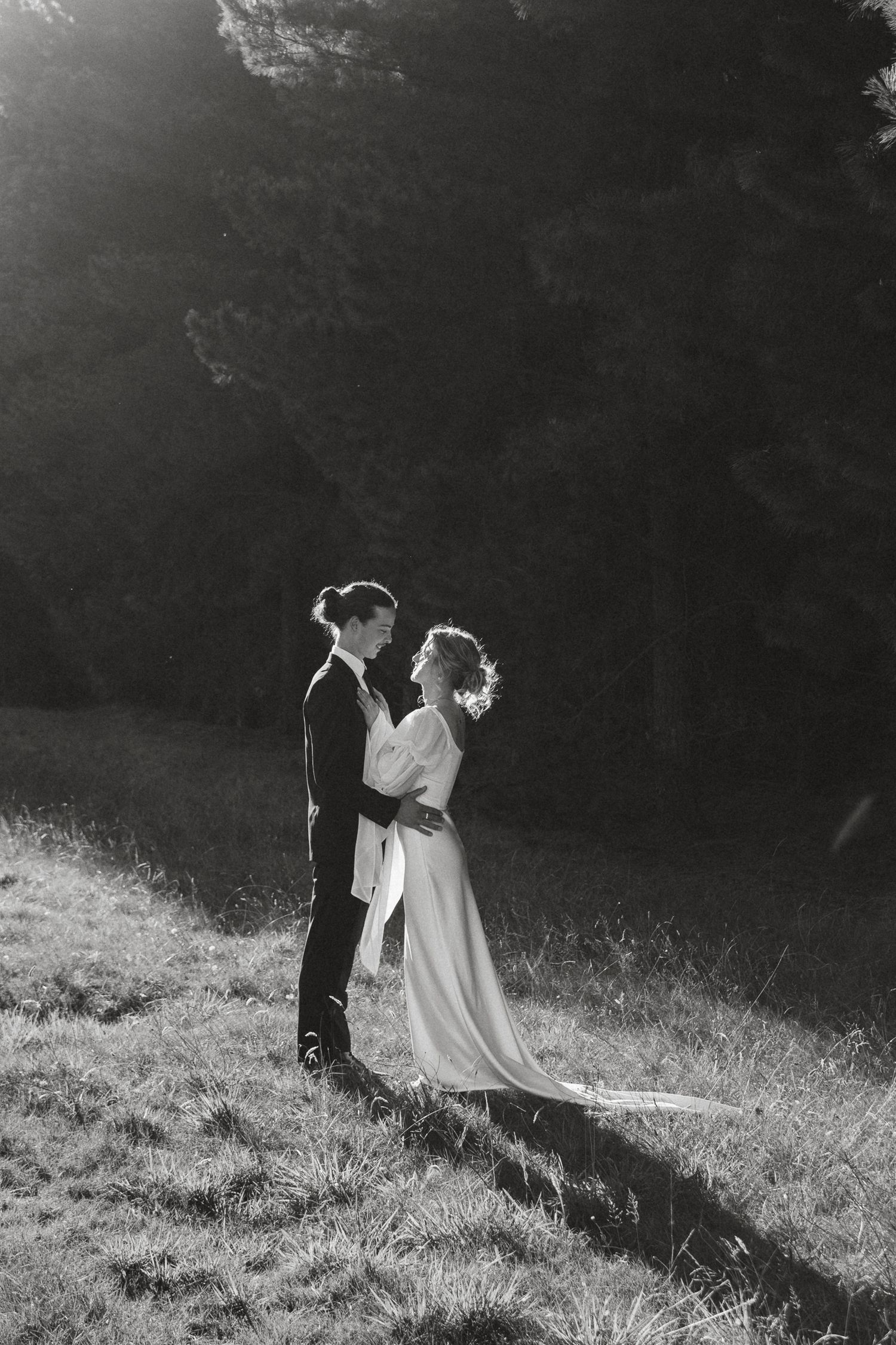 A couple in wedding attire stands together on a sunlit hillside in an artistic black and white photograph.