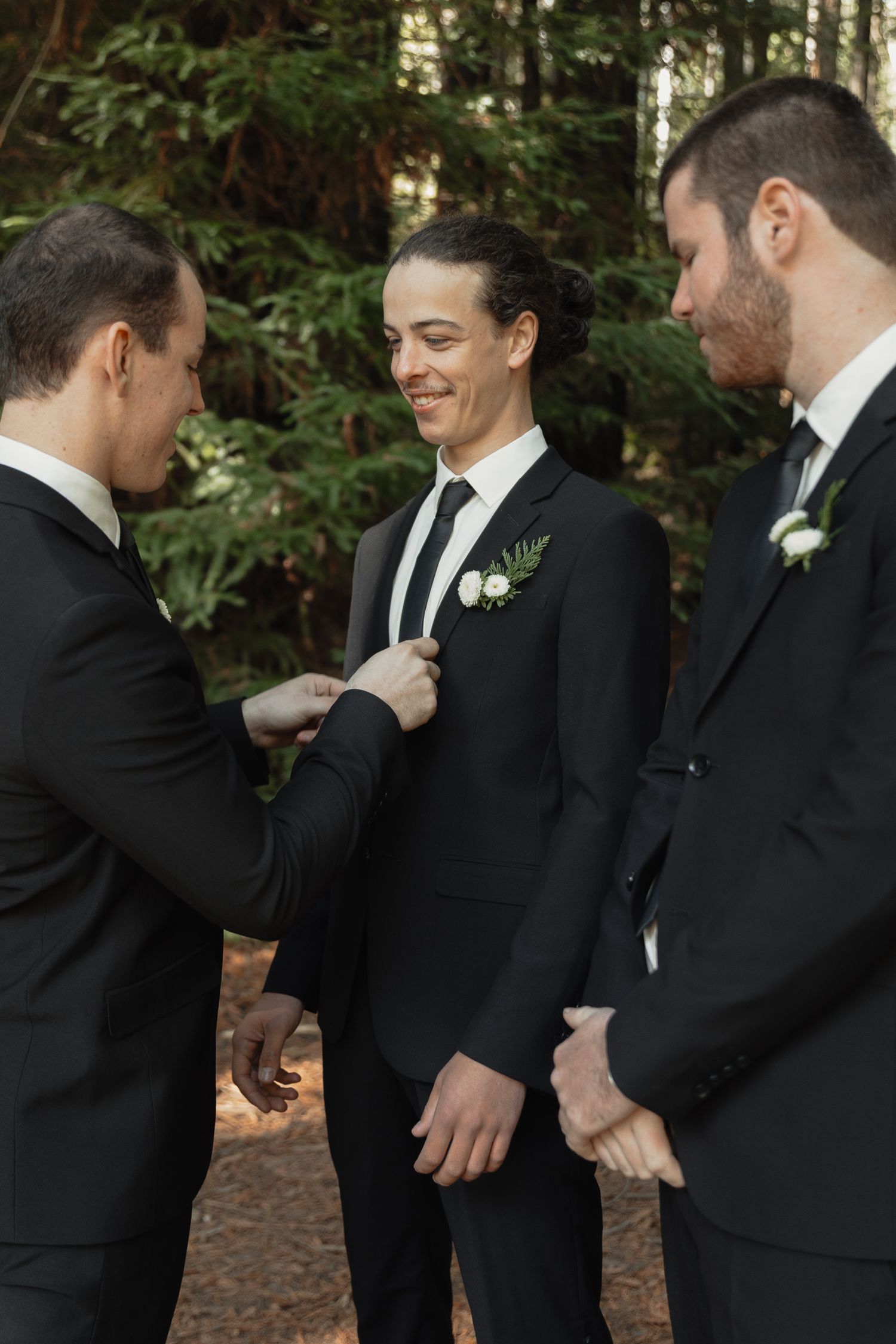 Groomsmen share a candid moment in black suits with floral boutonnieres against a forest backdrop.