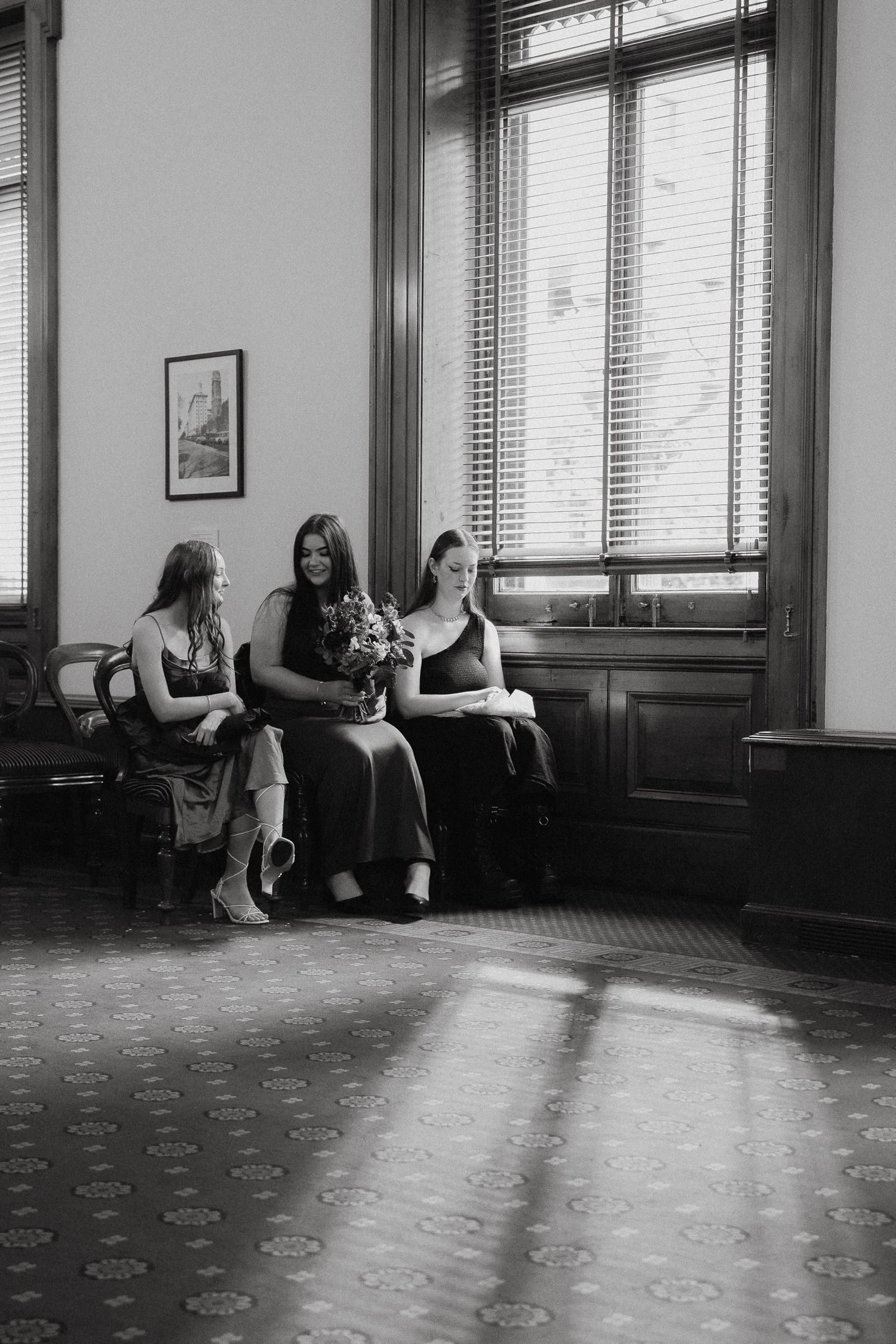 Three women sit on a bench in a bright room with tall windows, captured in black and white photography.