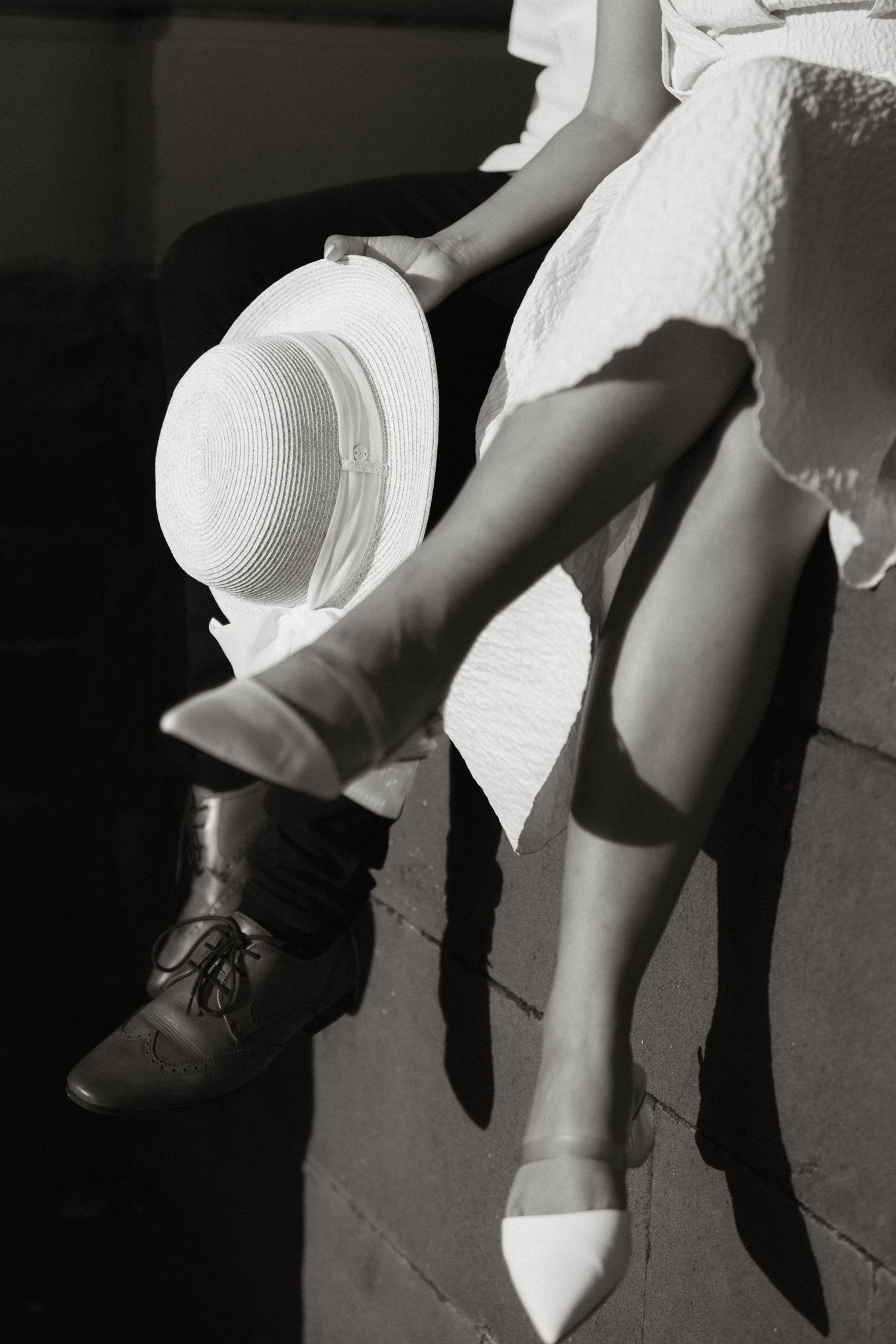 Black and white photo showing legs in contrasting shoes - one in dark oxford and one in white flat - with a white hat.