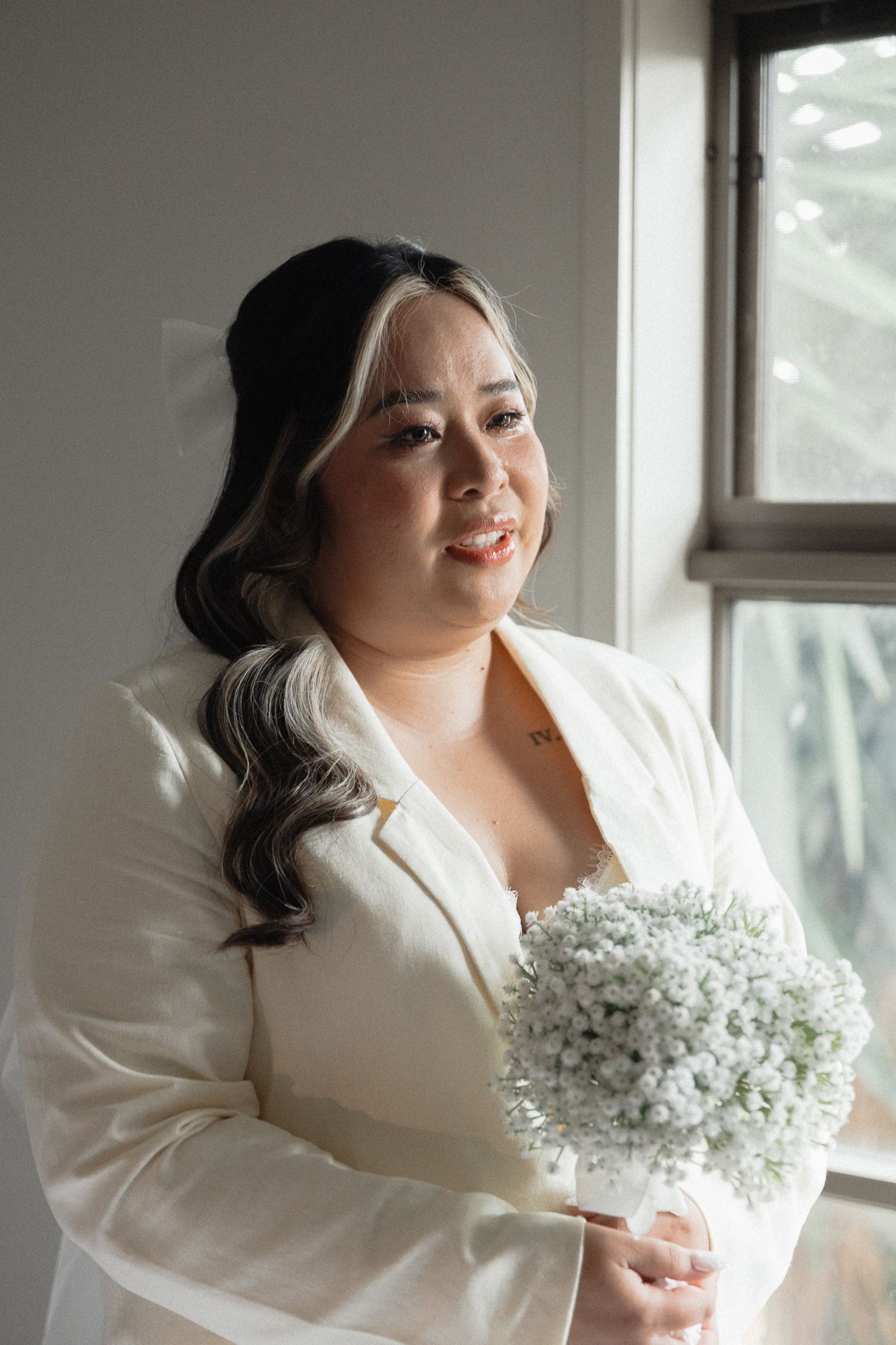 A person in a white blazer holds a delicate bouquet of baby's breath flowers while standing by a window.