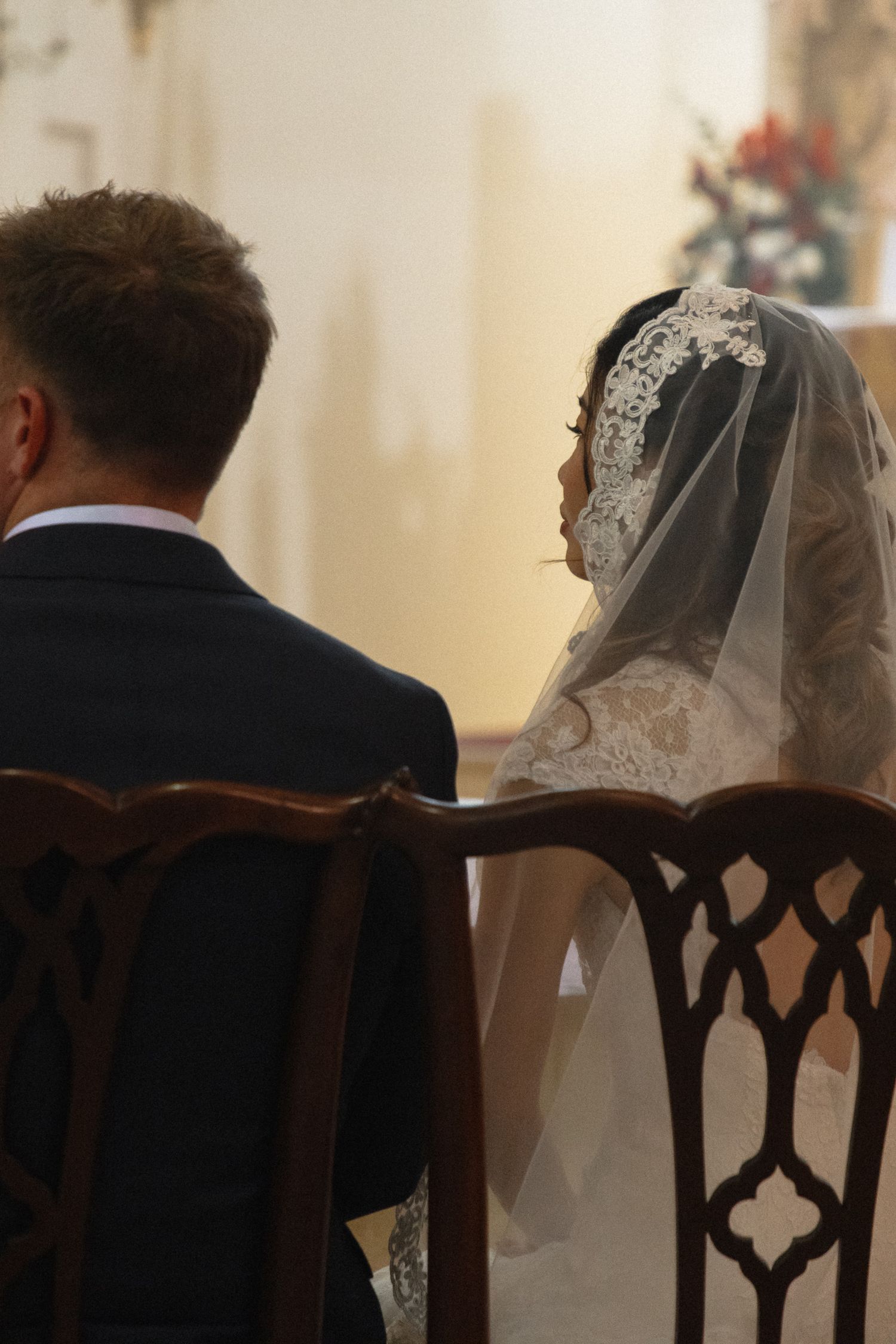 A bride and groom sit side by side in ornate wooden chairs during their wedding ceremony at a church.
