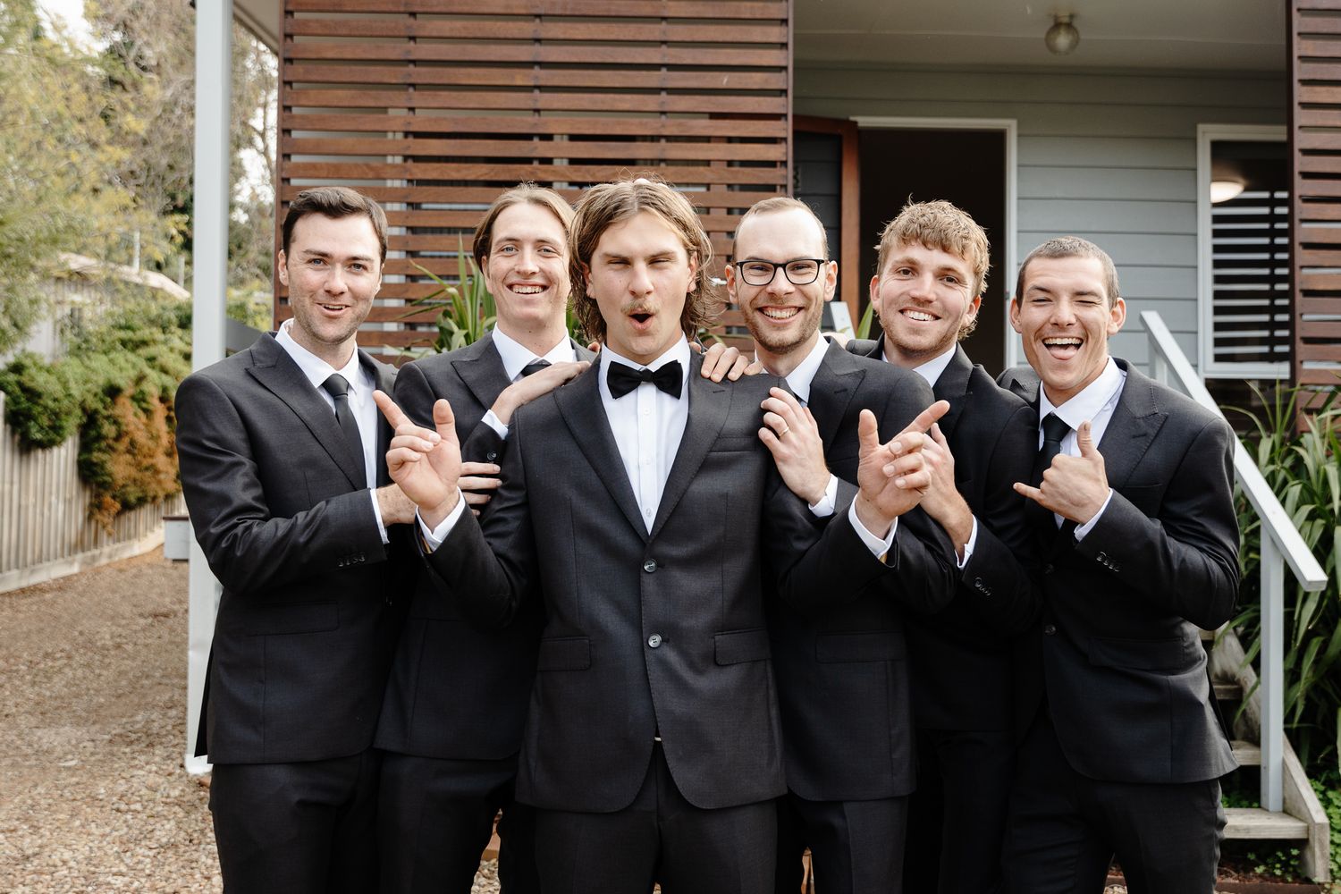 A group of groomsmen in black tuxedos and bow ties pose together smiling outside a house entrance.