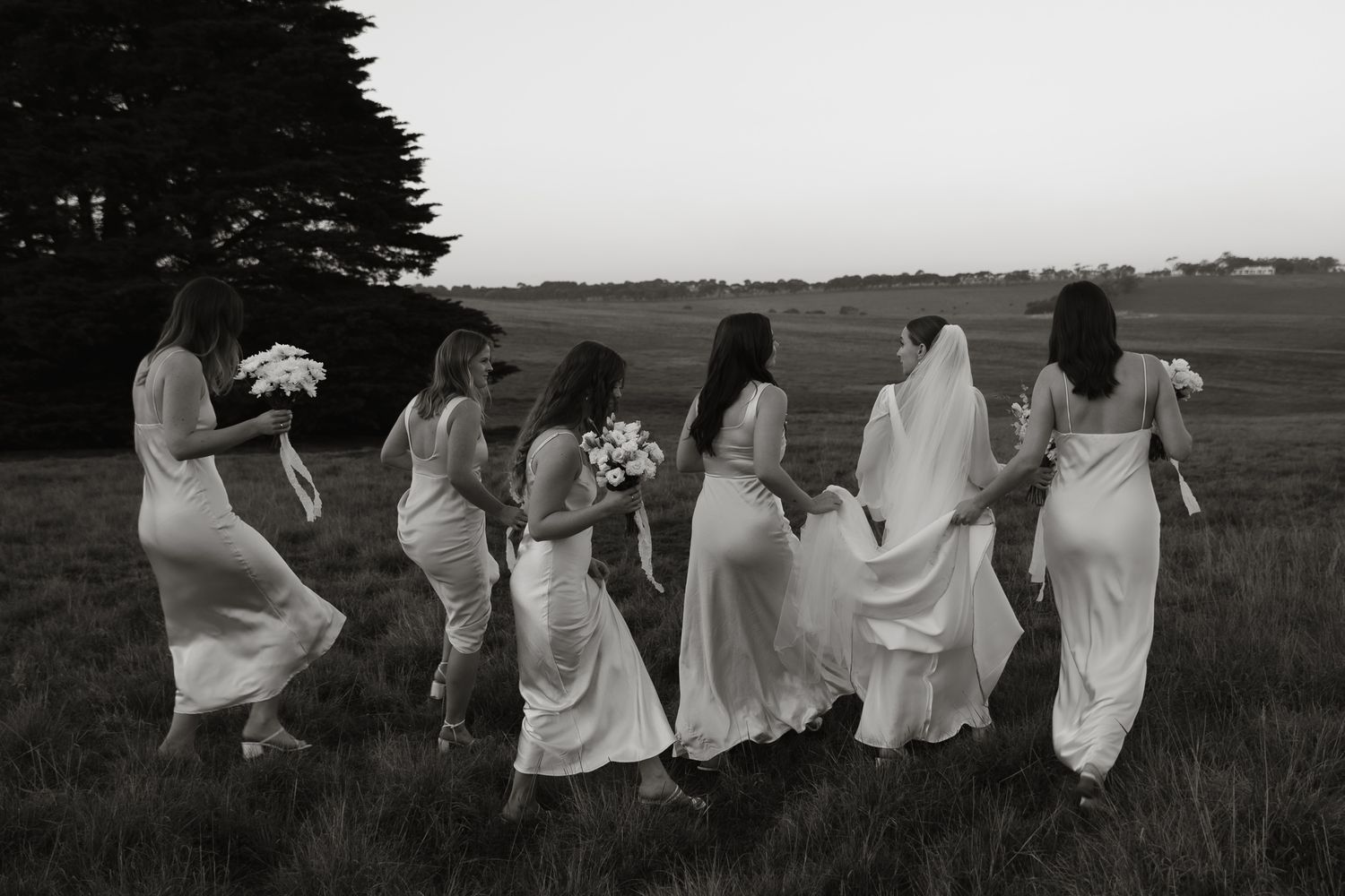 Group of bridesmaids in flowing white dresses running through a field at sunset.