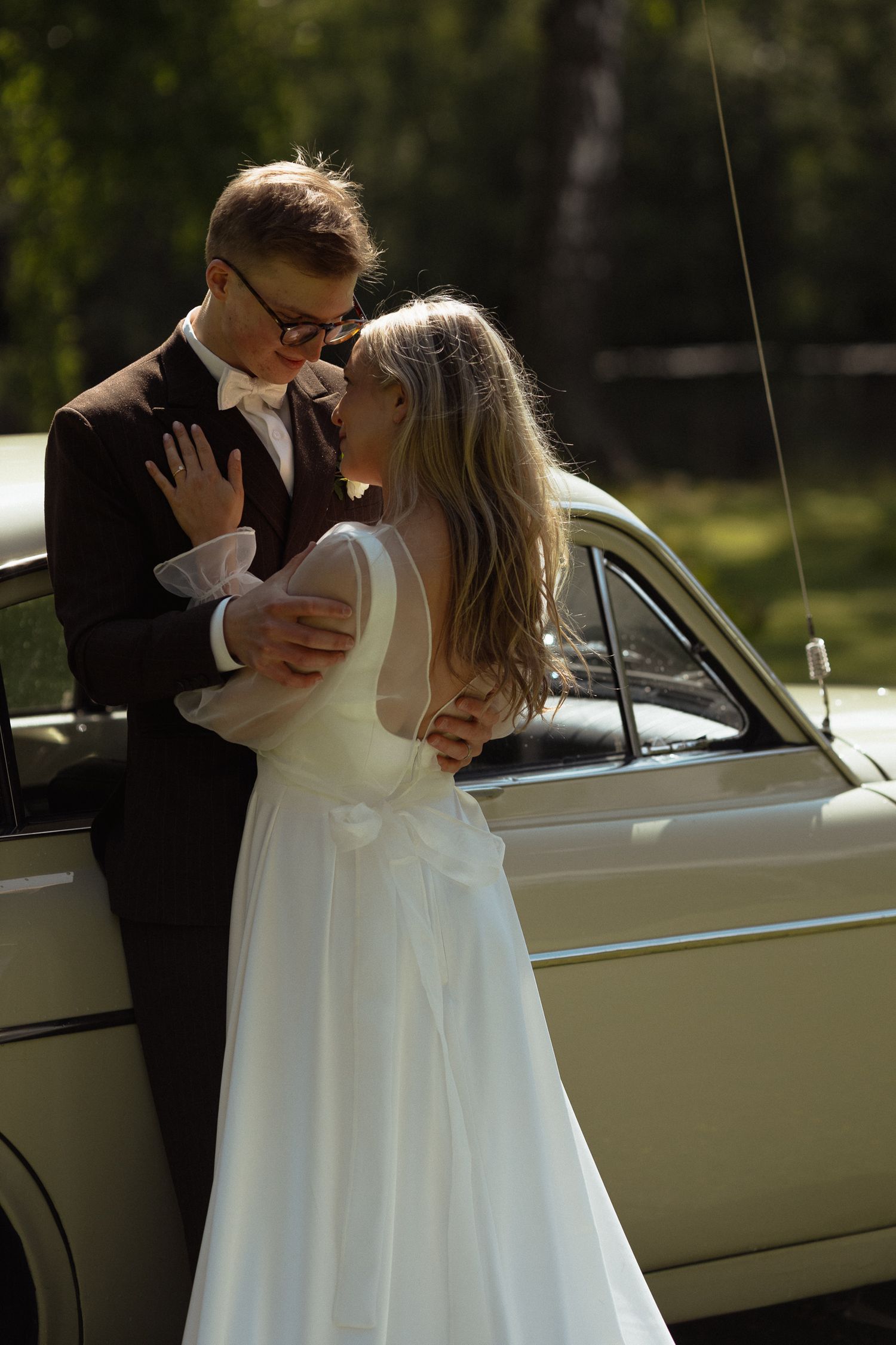 A romantic moment between a couple in wedding attire standing beside a vintage cream-colored car on their special day.