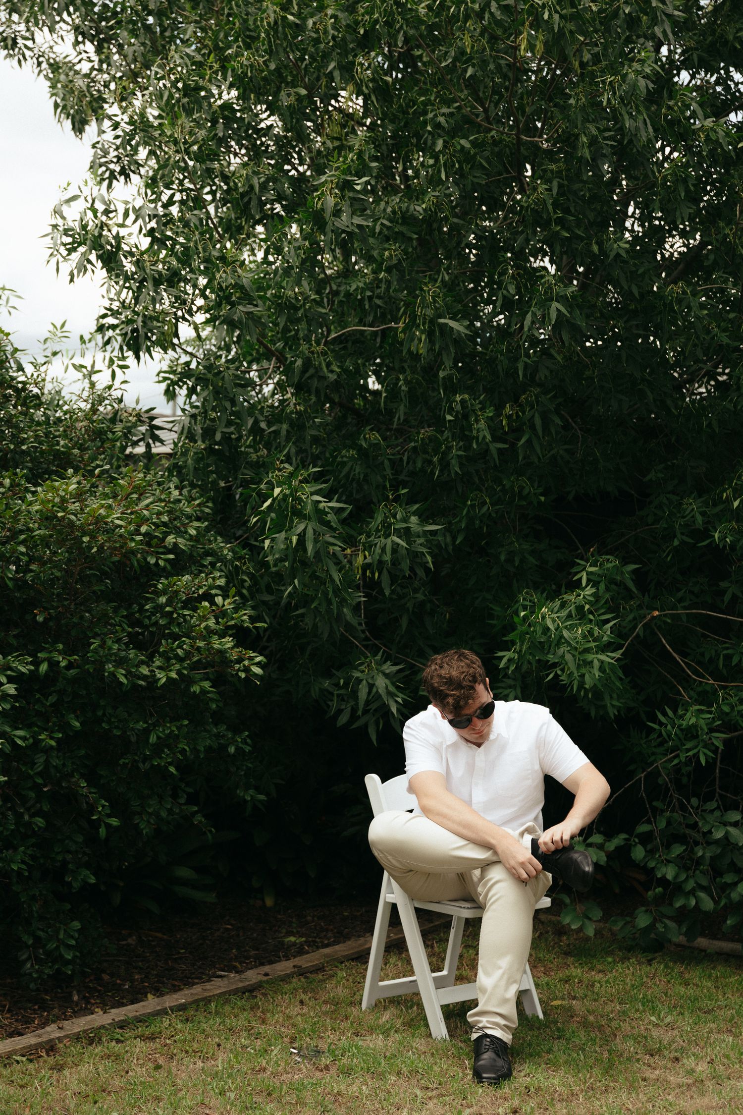 A figure in a white shirt and light pants sits on a white stool against a leafy green garden backdrop.