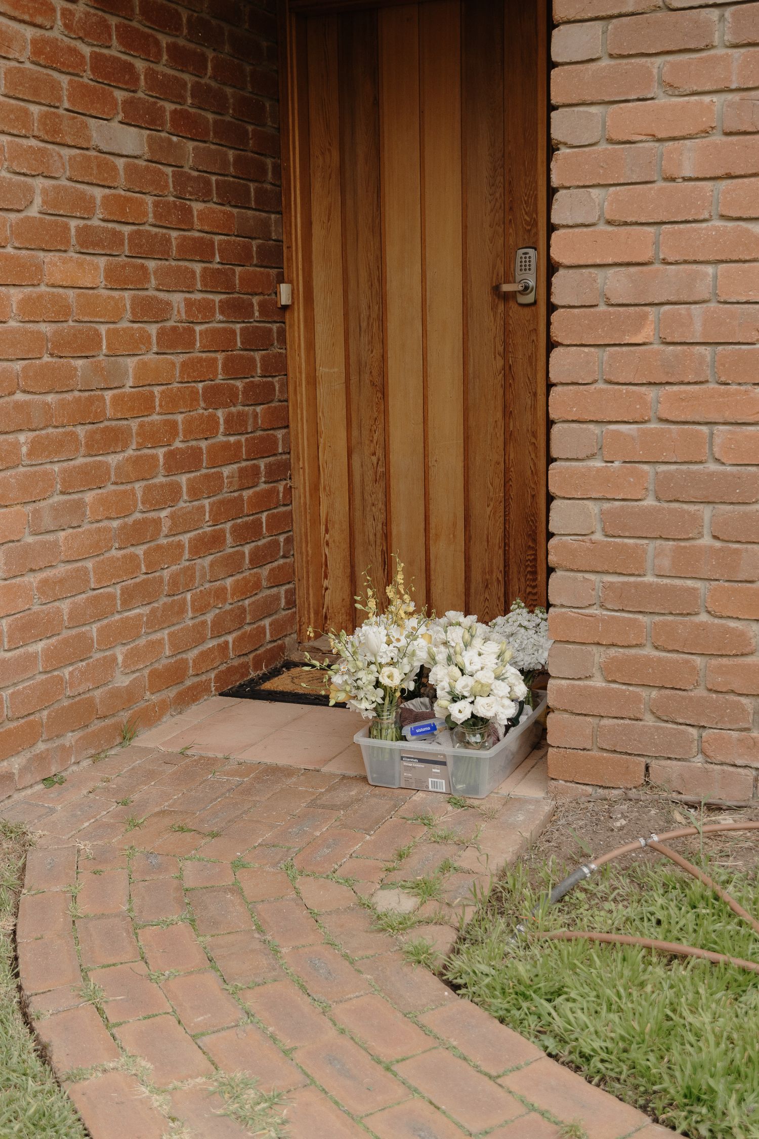 White flowers in a container sit at the entrance of a wooden door surrounded by red brick walls and pathway.