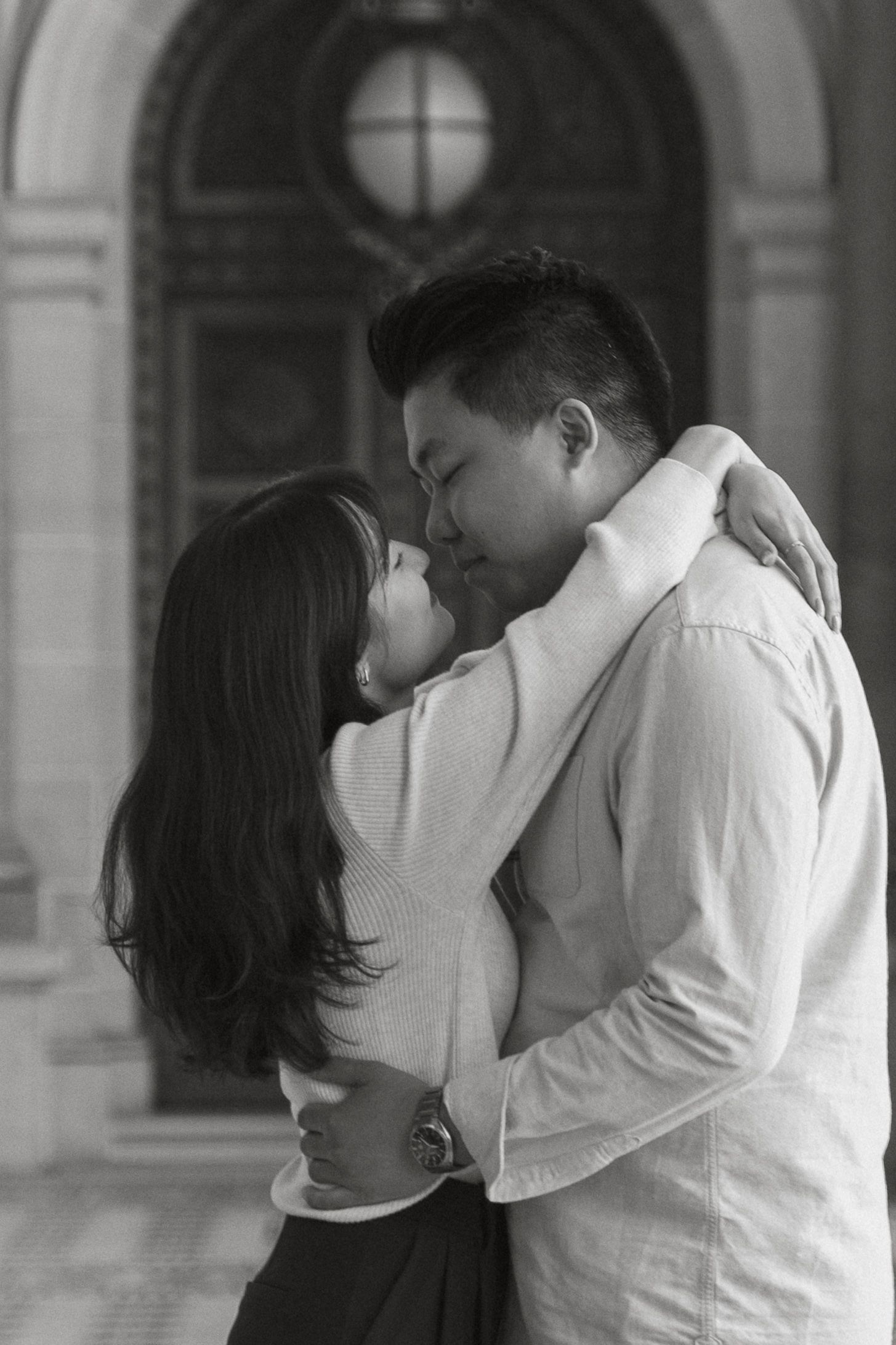 A romantic black and white portrait of a couple in white shirts embracing and sharing an intimate moment by an arched doorway.