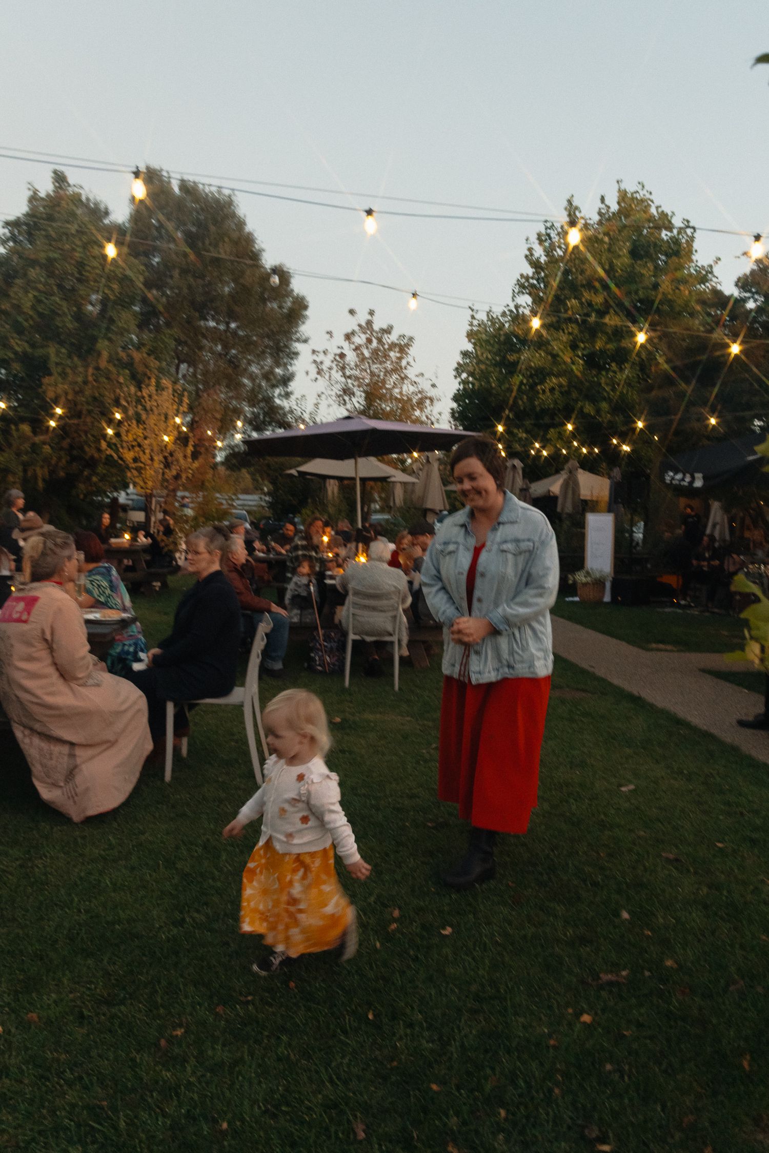 People gather at an outdoor evening party with string lights overhead and tables set up on a grassy lawn.